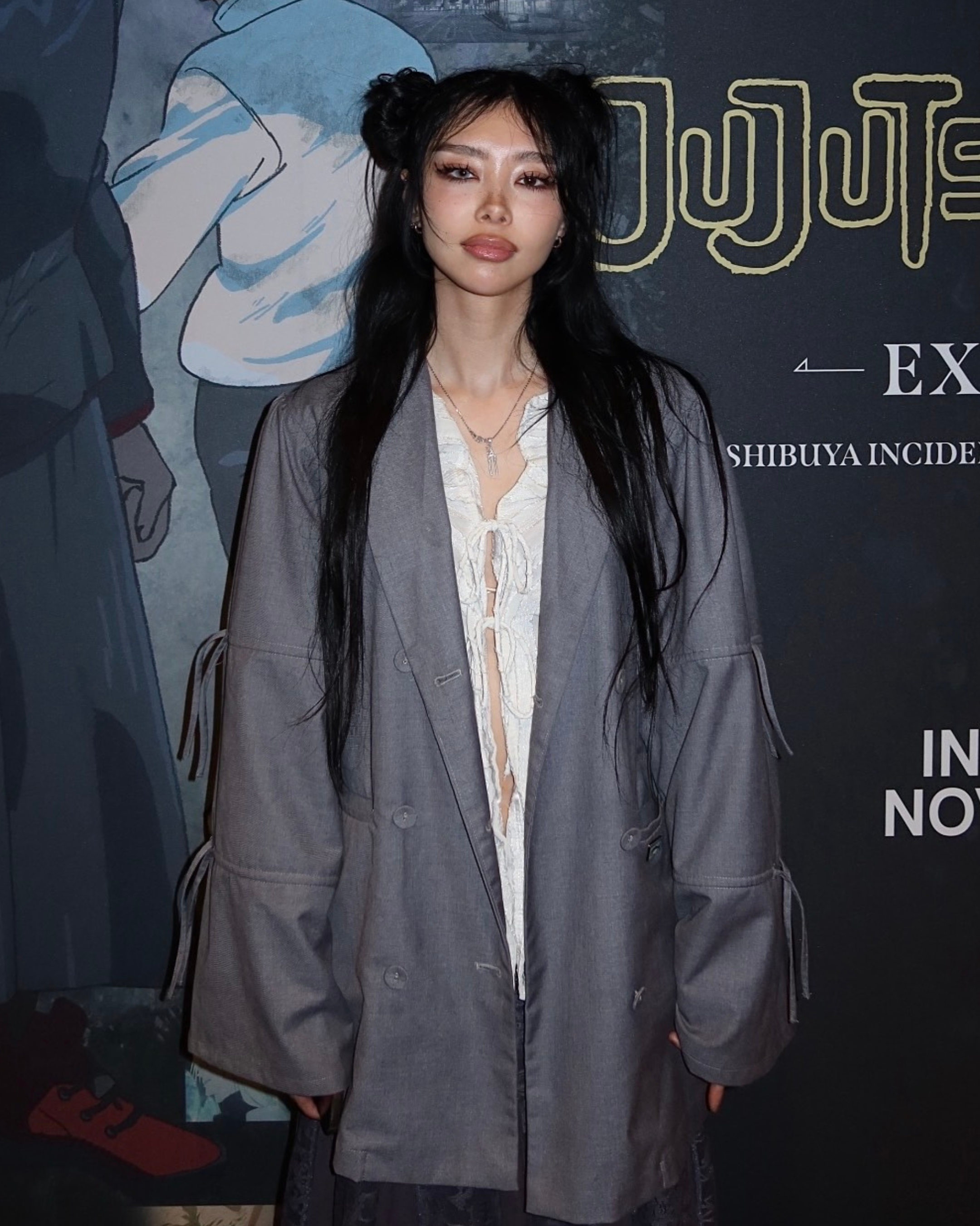 Young woman with long black hair styled in two buns, wearing a gray oversized blazer over a white lace-up blouse, standing in front of a promotional poster with illustrations and text.