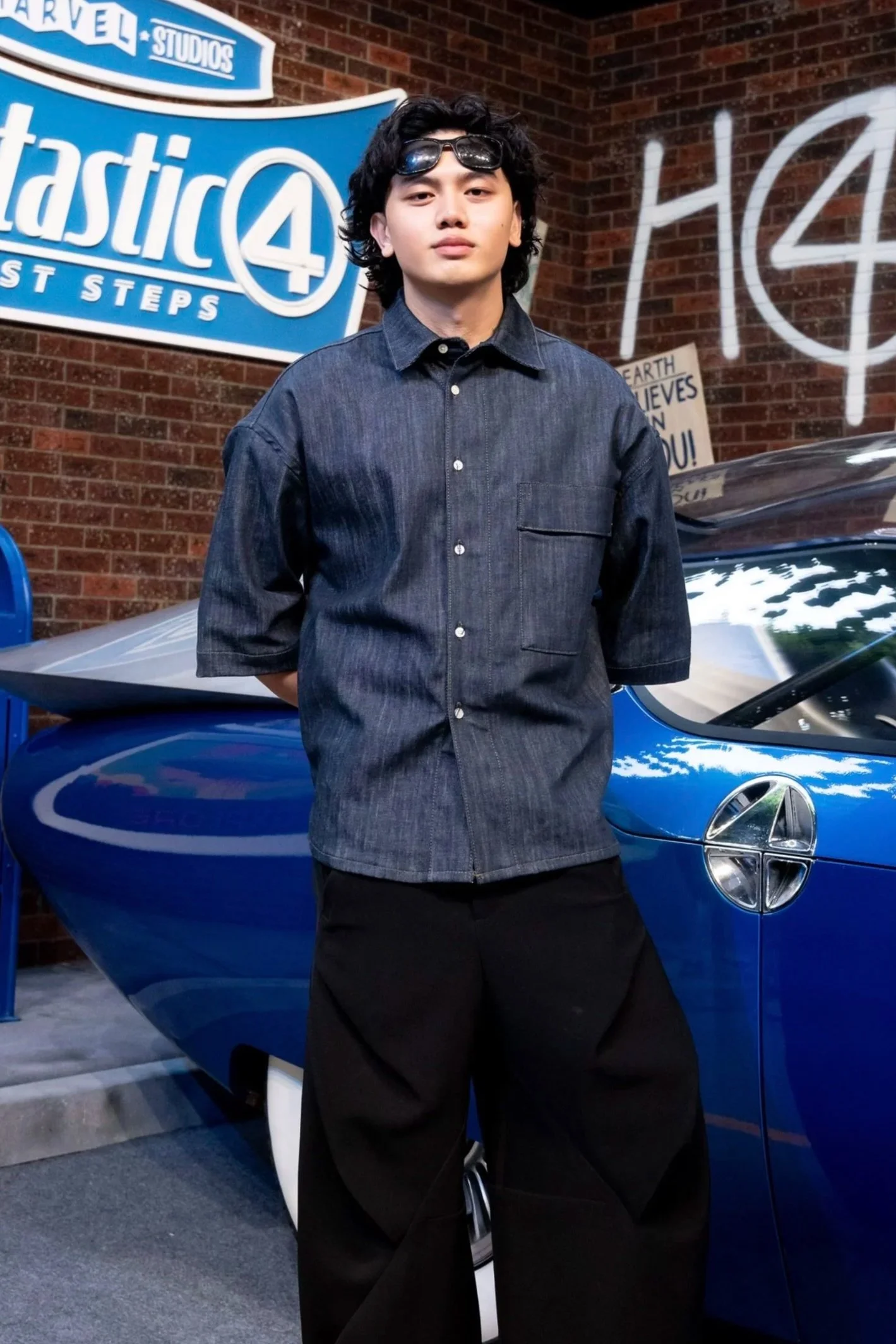 Young man with wavy black hair, wearing sunglasses on his head, a dark denim short-sleeve shirt, and black pants, standing in front of a blue sports car at Marvel Studios' Astic 4 sign.