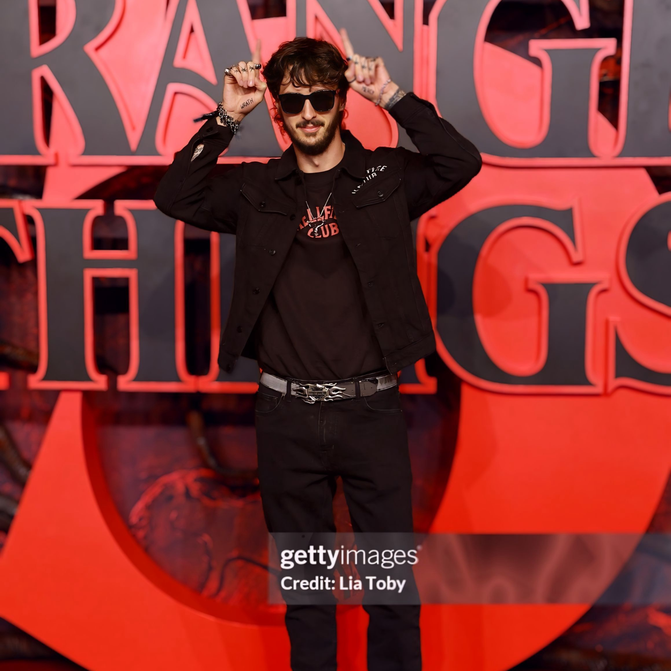 A man with brown hair and beard wearing sunglasses, a black jacket over a black T-shirt, and black pants with a decorative belt, standing in front of a red and black background with large stylized text.