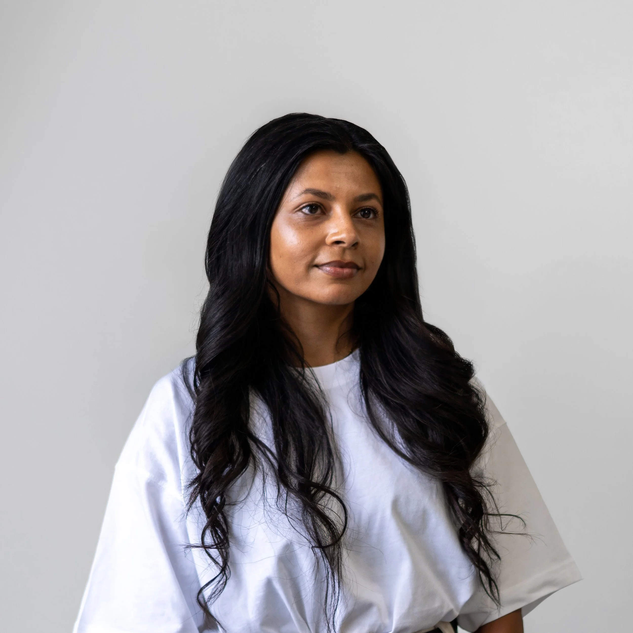 A woman with long, wavy black hair wearing a white shirt, standing against a plain, light-colored background.