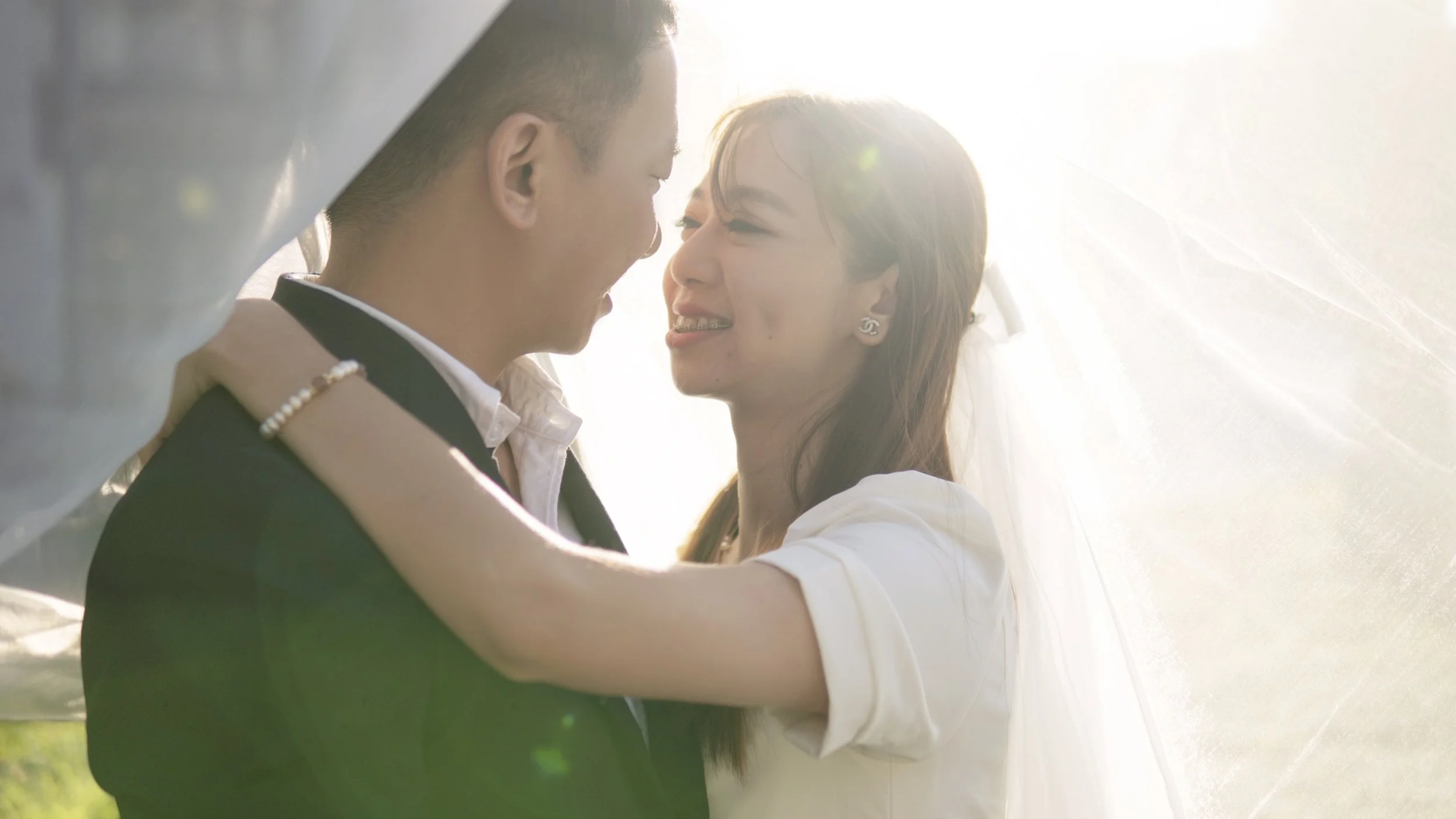 A happy couple is embracing and smiling under a sheer, white fabric during a sunny outdoor wedding, with sunlight creating a bright halo around them.
