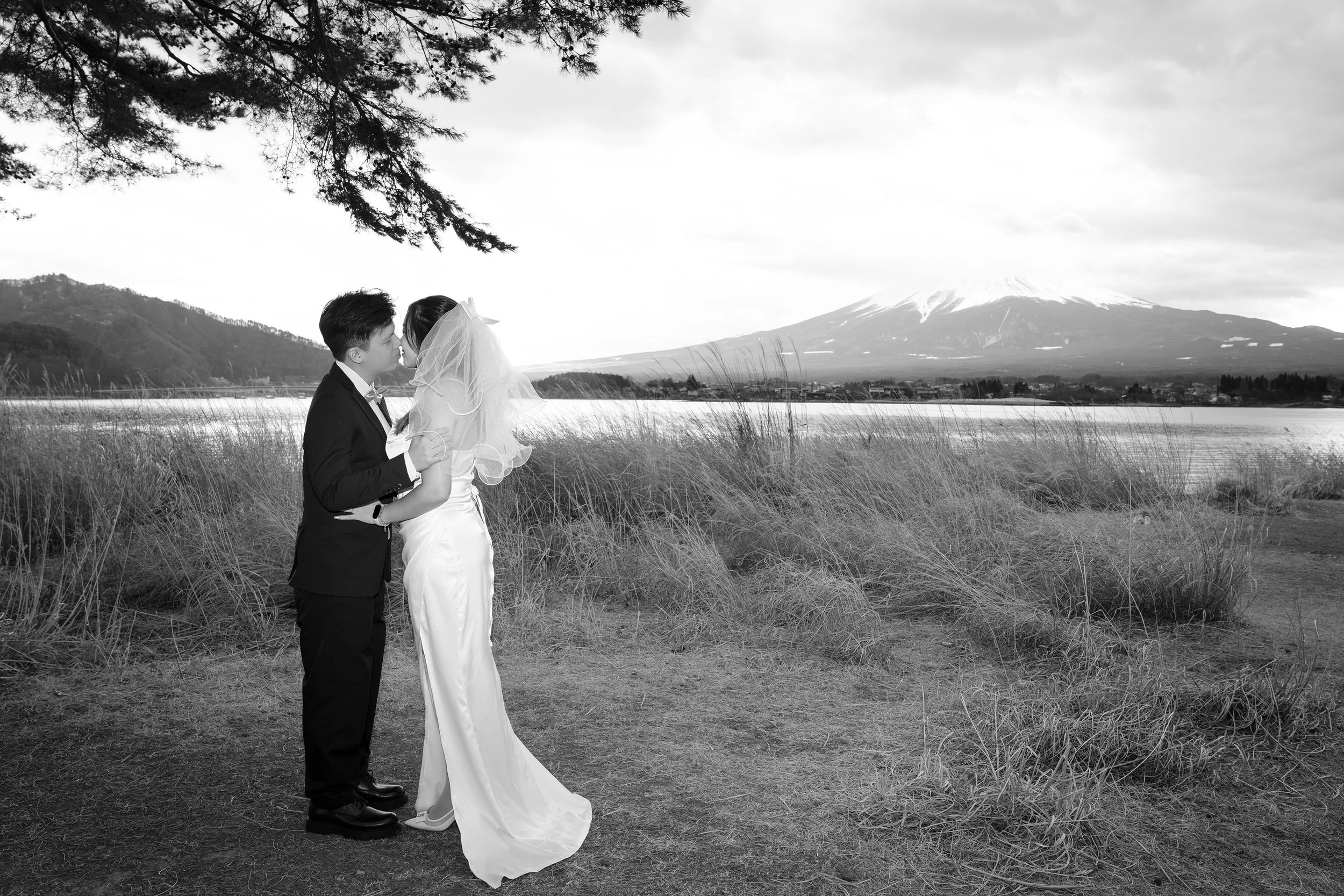 A wedding couple kissing outdoors near a lake with Mount Fuji in the background, black and white photo.