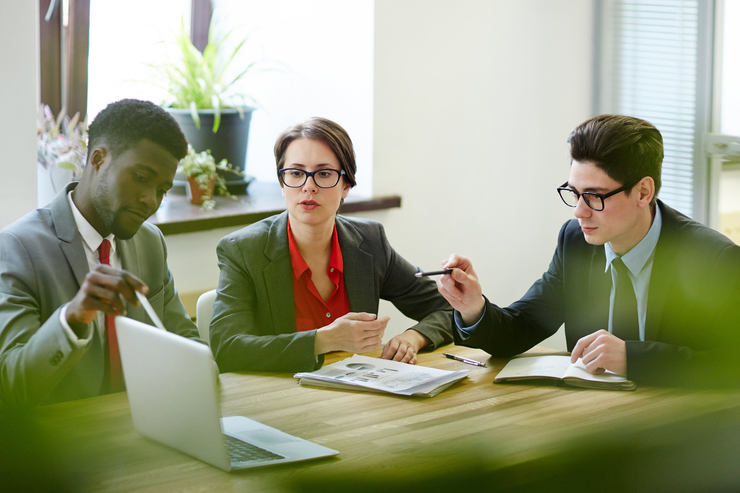 Three professionals in business attire and glasses sitting at a wooden conference table with laptops, notebooks, and papers, engaged in a discussion in a bright office with large windows and potted plants in the background.