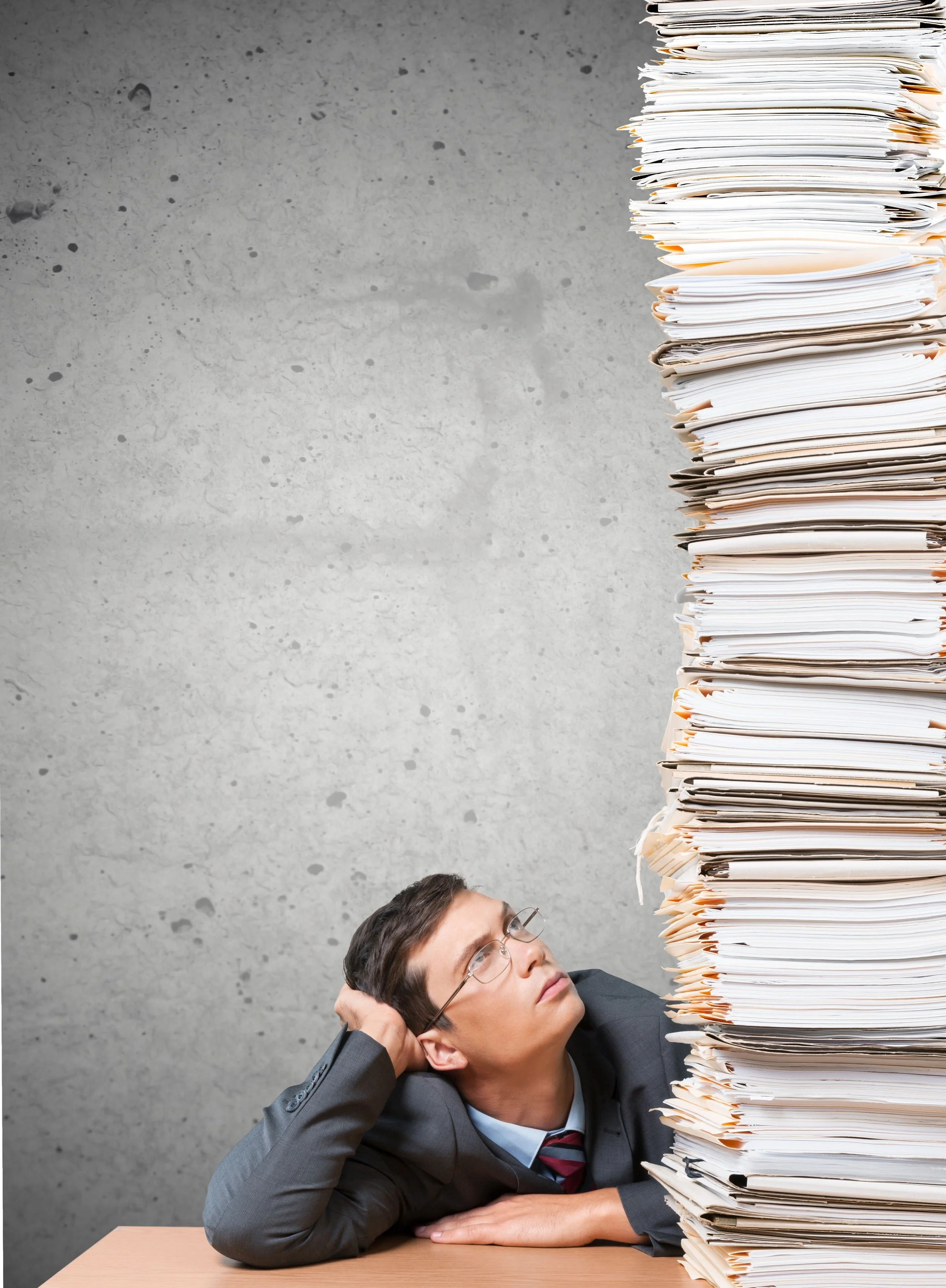 A man in business attire resting his head on his hand at a desk, looking up at a large stack of disorganized paperwork and folders.