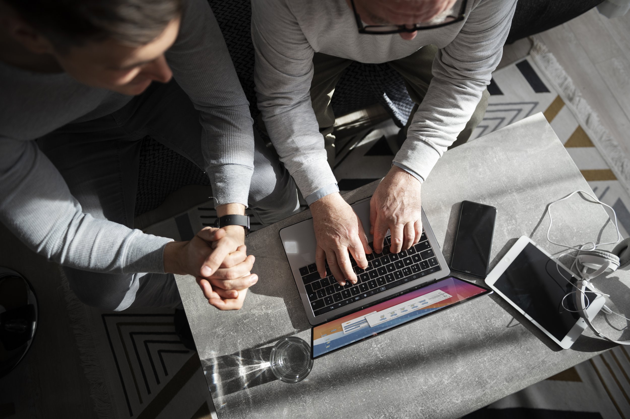 Two people sitting at a table working on a laptop, with a smartphone, tablet, and headphones nearby, and sunlight casting shadows on the table.