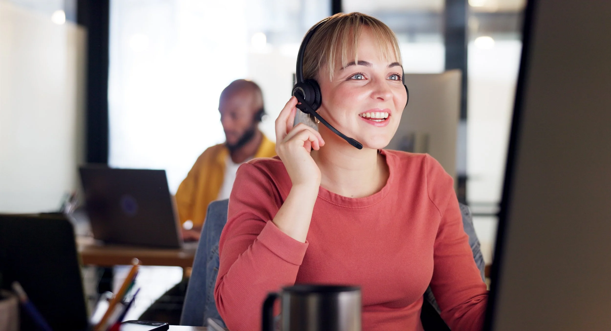 A smiling woman wearing a headset working at a computer in an office with a male colleague in the background.