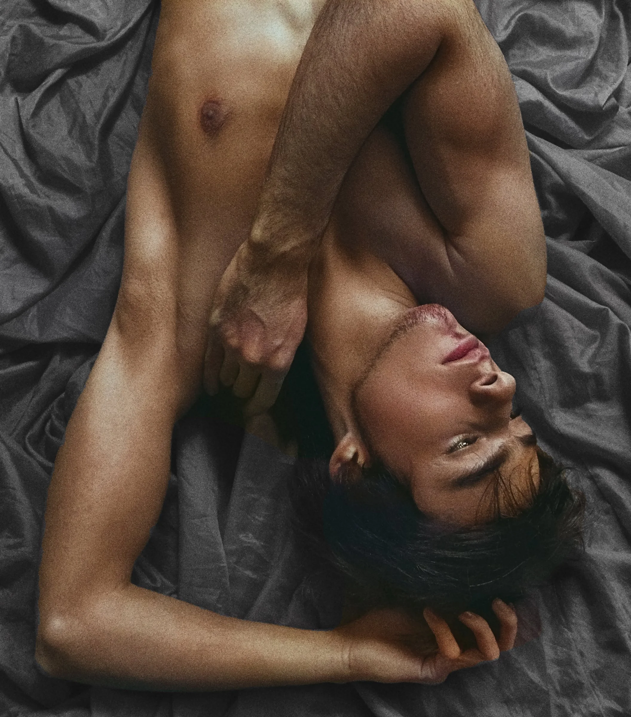 A woman lying on a dark gray sheet, with a man lying on top of her, partially unclothed. The woman has dark hair and appears to be relaxed.