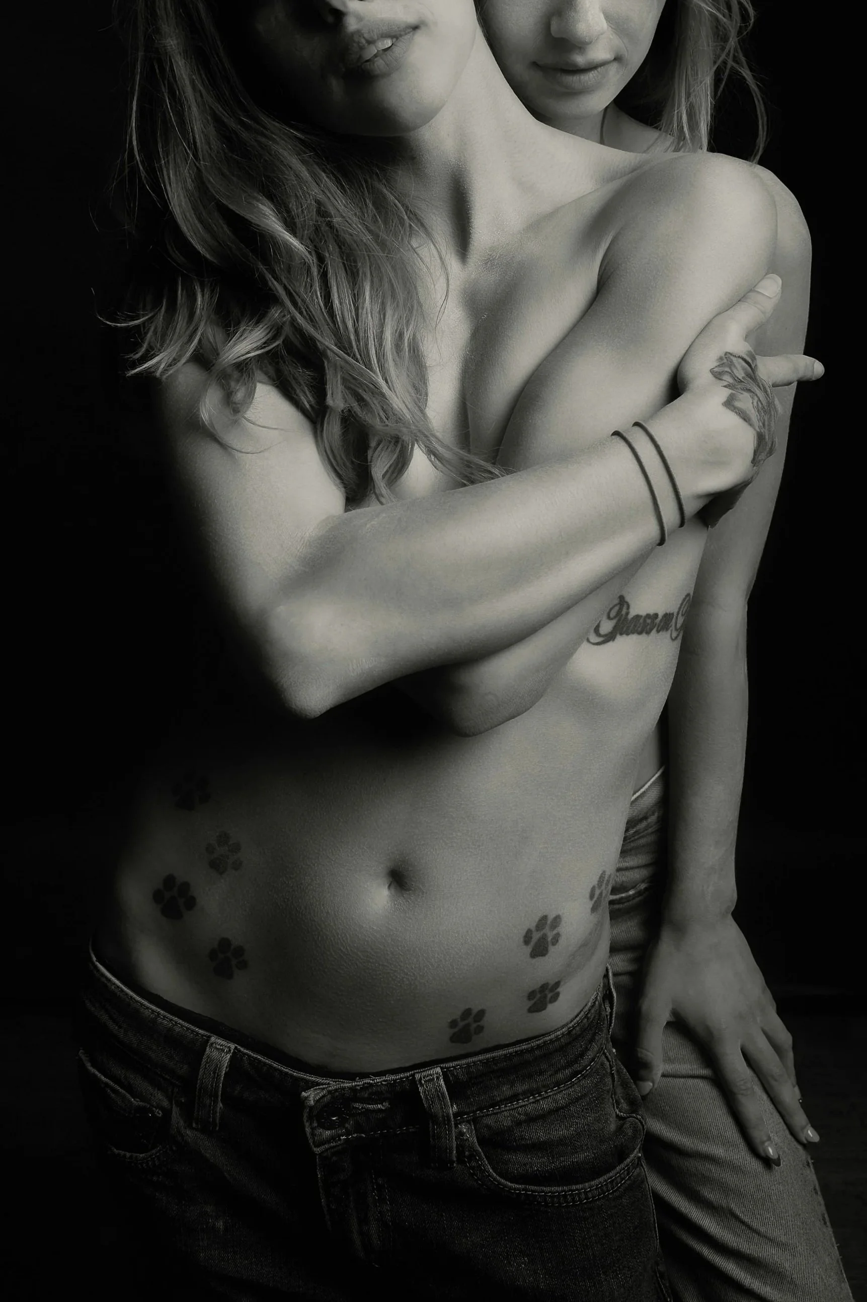 A black and white photo of two women, one with long wavy hair and tattoos, partially covering her chest, and the other with shorter hair, standing behind her, both with calm expressions.