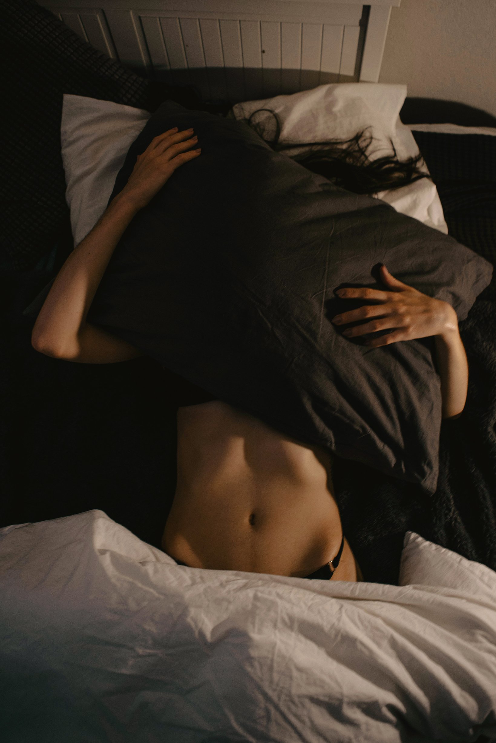 Person lying on a bed, partially covered with a black blanket, with visible bare midriff and hand resting on the blanket.