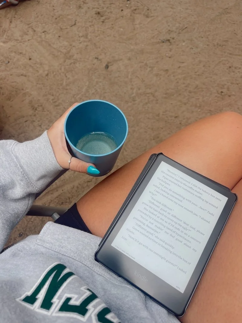 Person sitzt auf einem Strand, liest ein E-Book auf einem Kindle und hält ein Glas Wasser in der Hand.