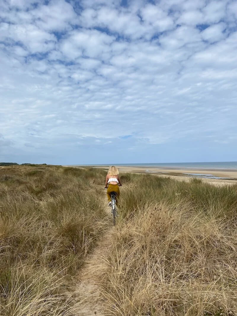 Eine Frau radelt durch Gräser auf einem Sandweg an der Küste entlang, im Hintergrund Meer und Himmel mit Wolken.