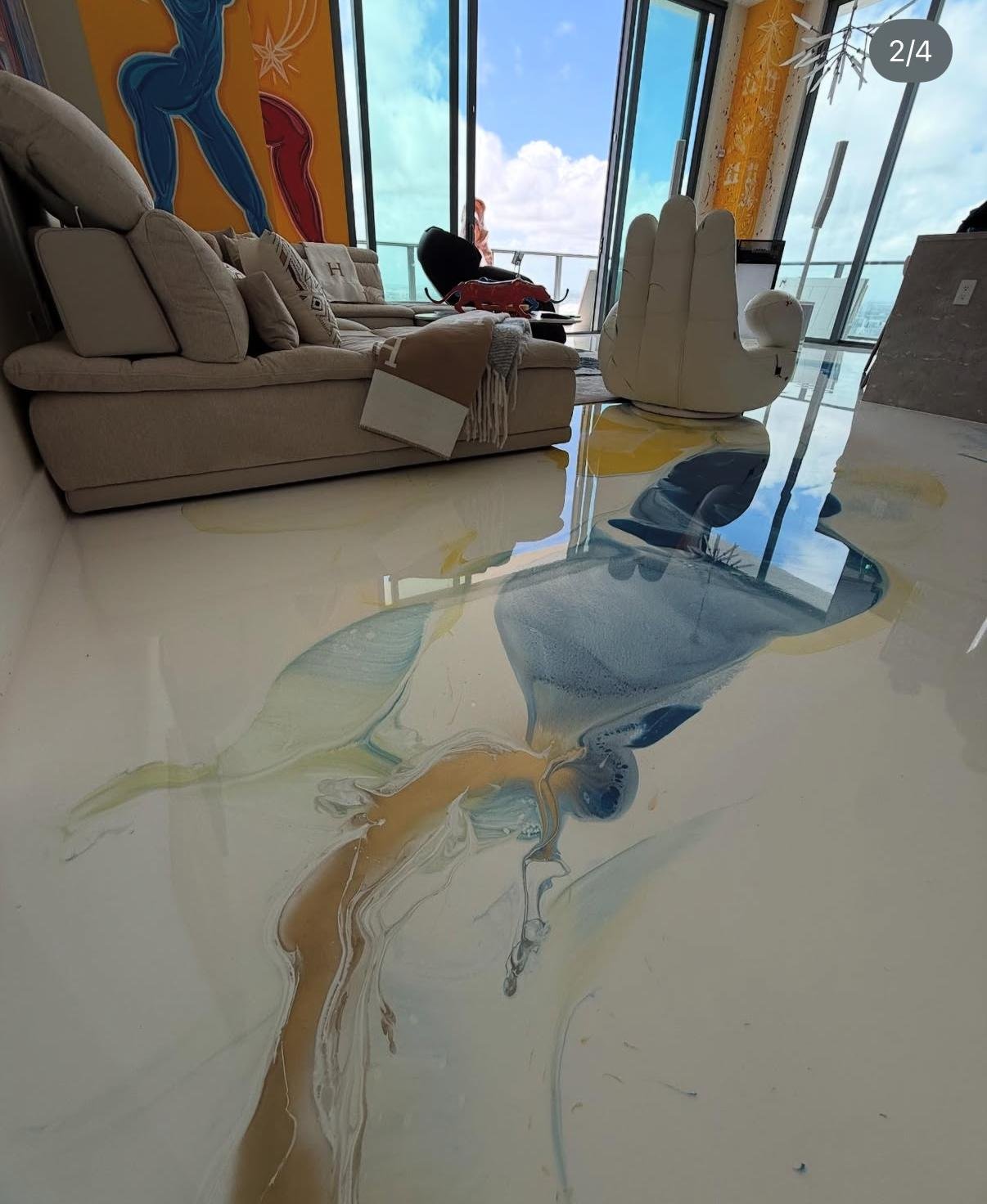 Modern living room with a large beige sectional sofa, a unique white hand-shaped chair, and glass walls showing a cloudy sky outside. The polished floor has colorful abstract paint patterns.