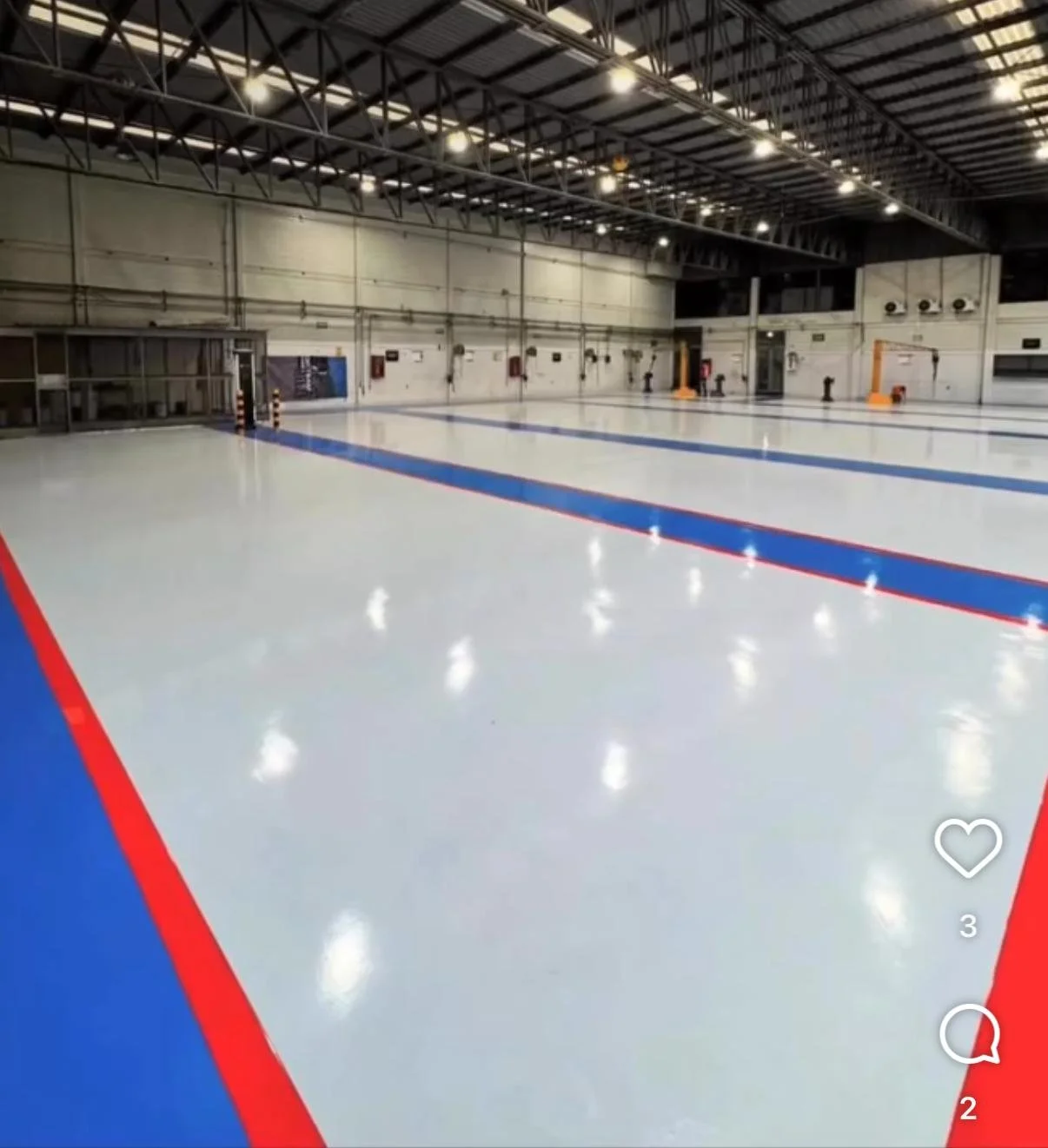 Indoor ice skating rink with freshly mopped ice, red and blue lane markings, and bright overhead lighting.