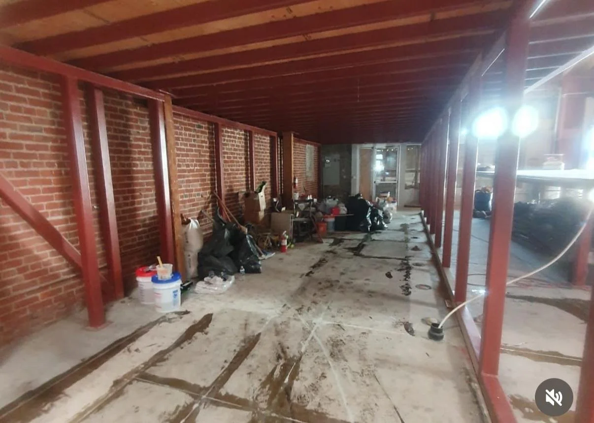 Incomplete room under construction with exposed brick walls, metal studs, scattered construction supplies, and a concrete floor with patches of wet concrete.