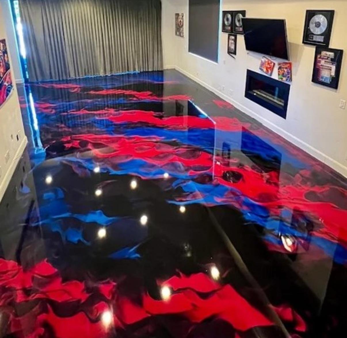 Living room with a glass table, television, framed gold records, and memorabilia on the walls. The table has a colorful abstract design resembling swirling red and blue flames, giving it a fiery appearance.