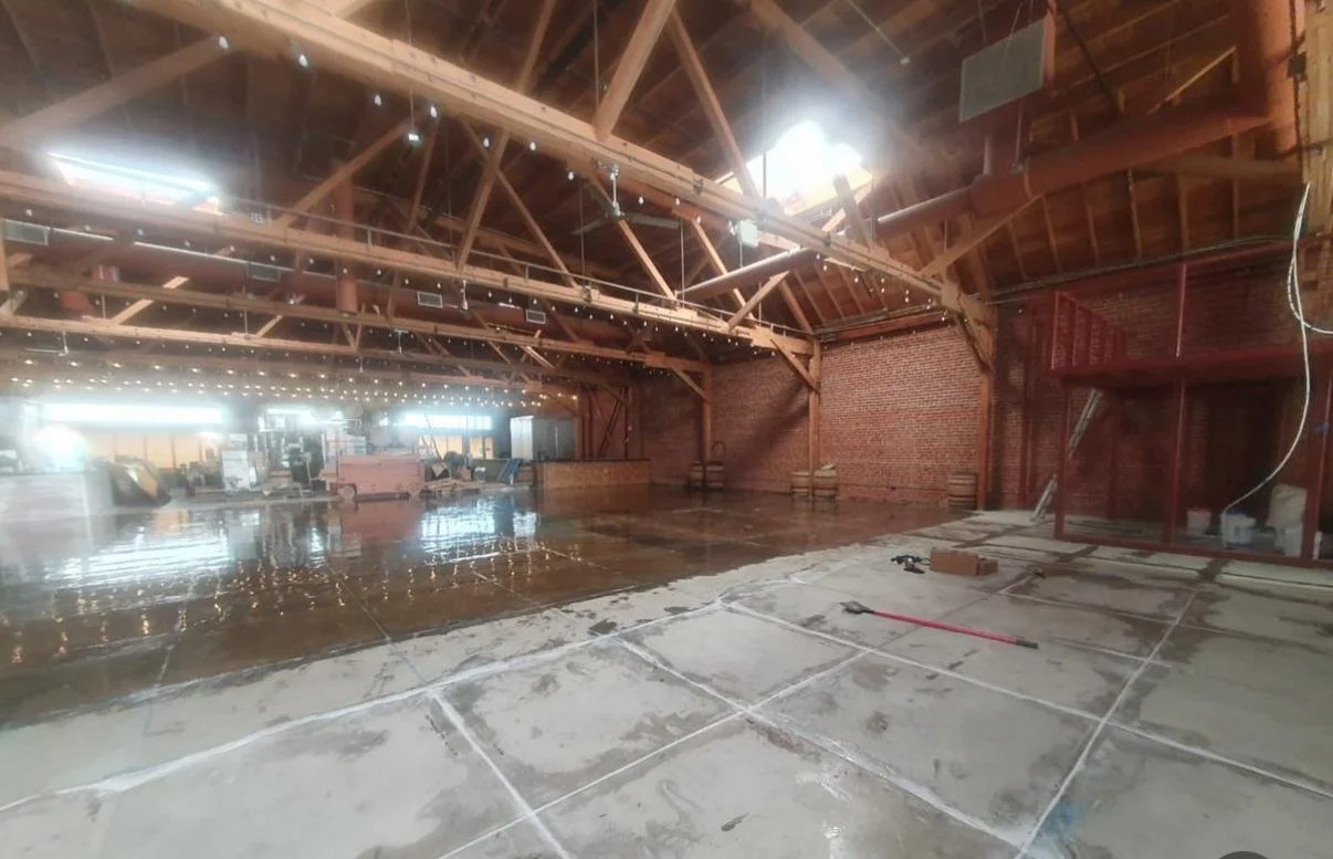 Construction of a wooden ceiling and brick wall in a large indoor space, with flooring being prepared and construction tools scattered around.