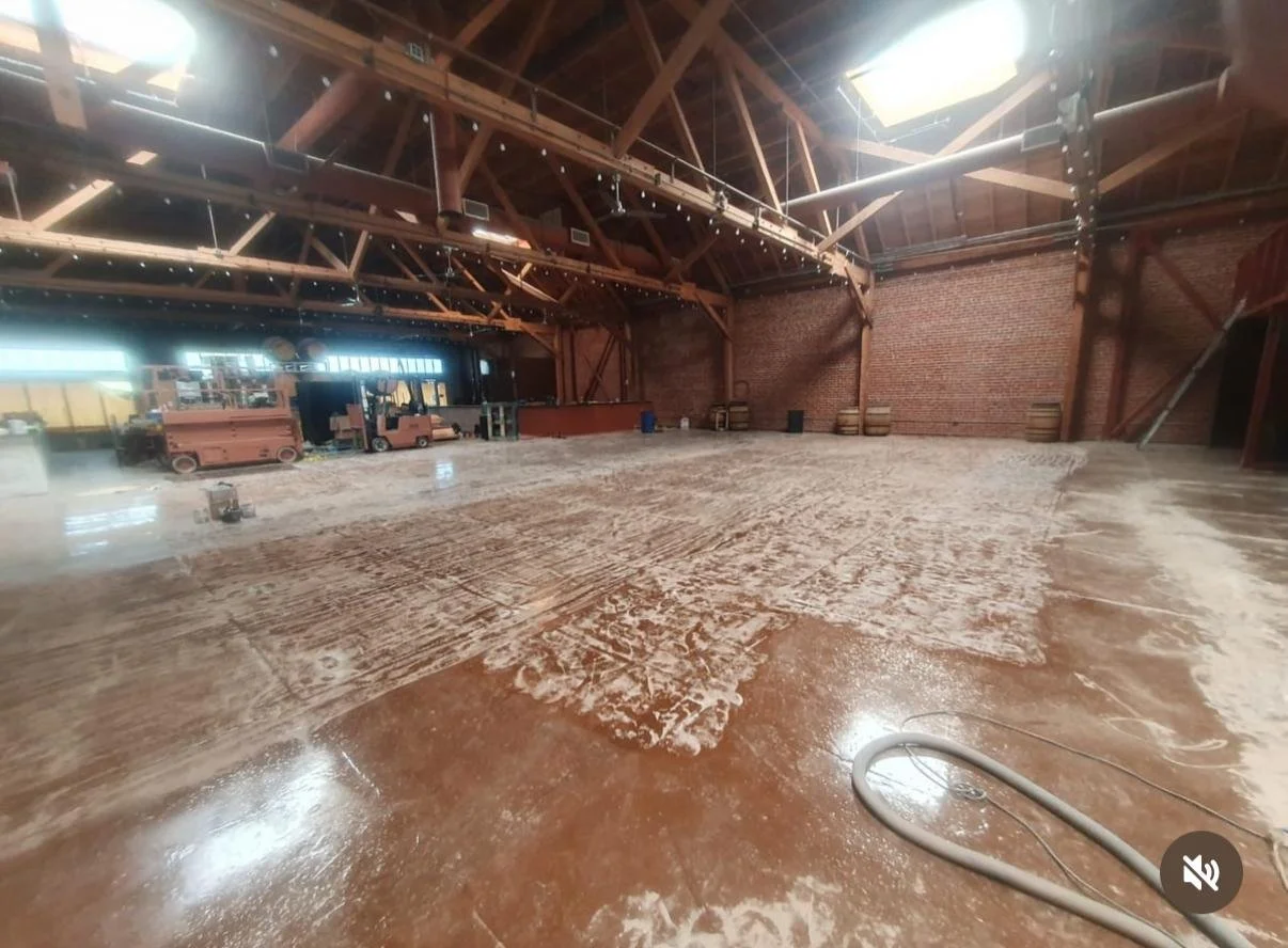 Empty warehouse space with a wet floor and construction materials, exposed wooden beams, and brick walls in the background.