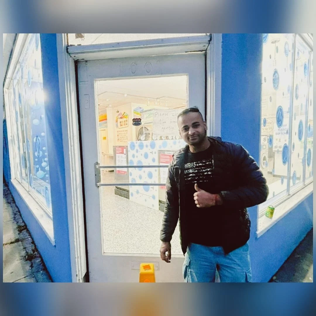 A man standing outside a blue building with large windows, giving a thumbs-up gesture.