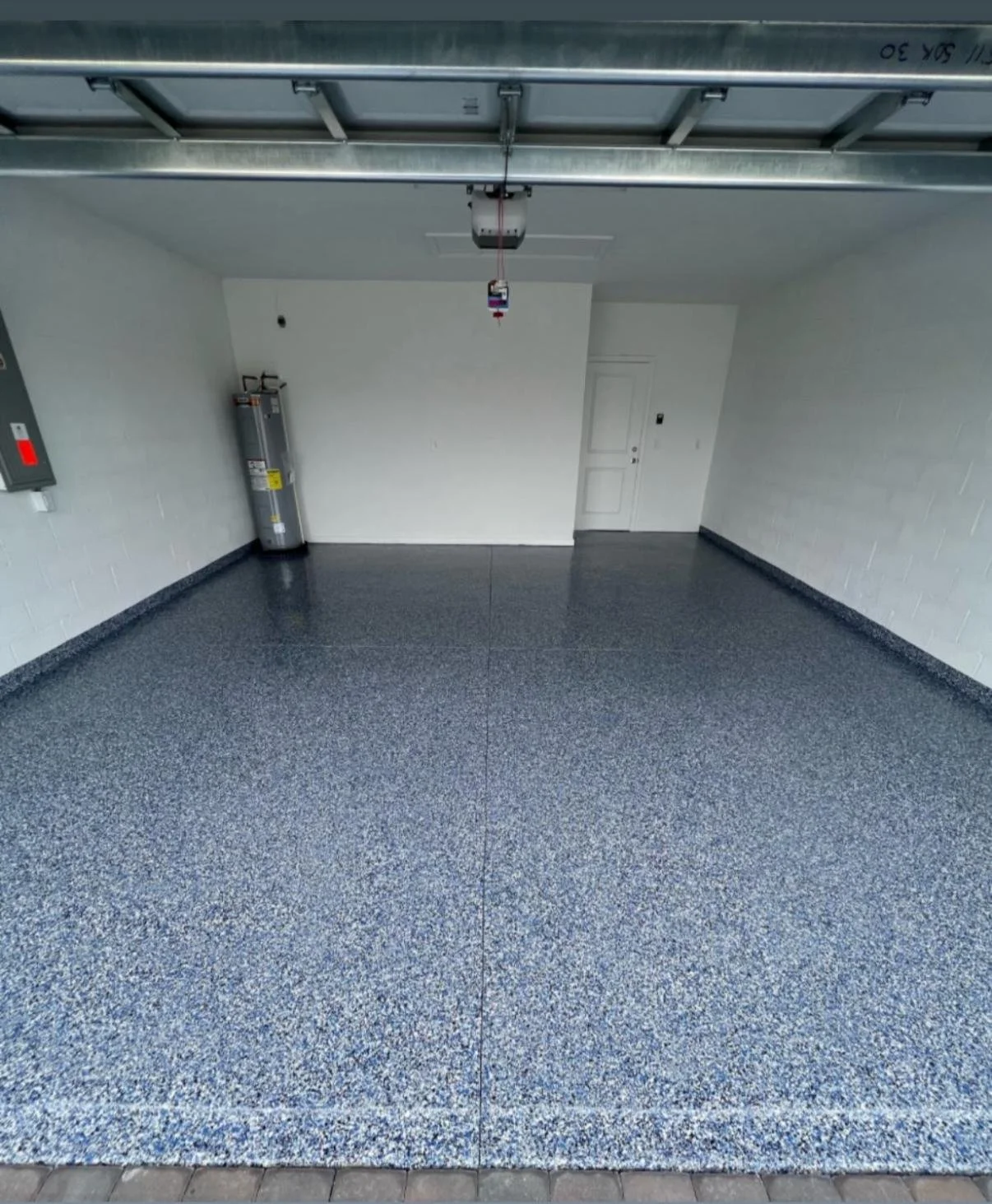 Empty garage with speckled blue-gray epoxy floor, white brick walls, a closed white door, water heater in the corner, and garage door opener above.