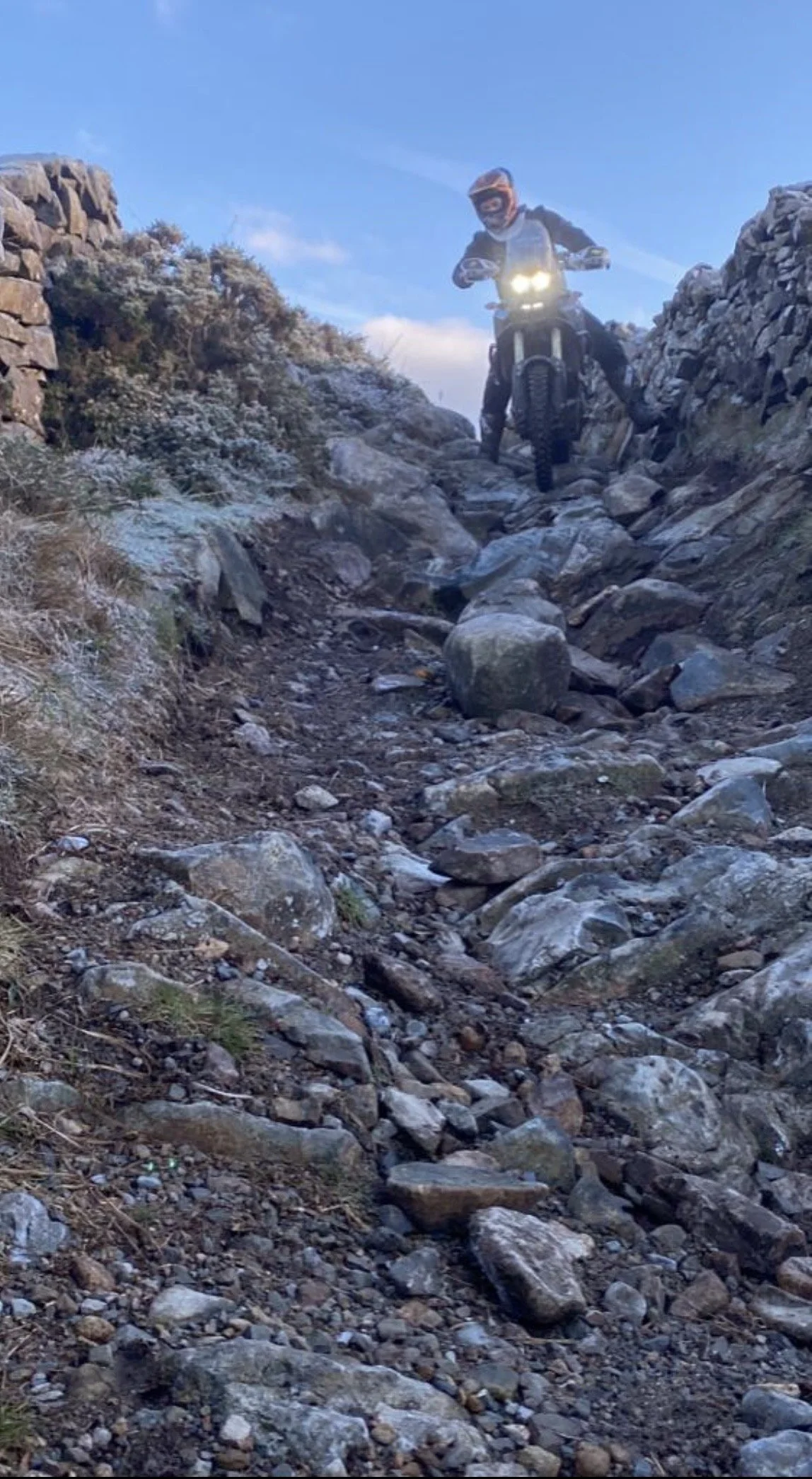 A person riding a dirt bike navigating a rocky trail outdoors, with stone walls on either side and a blue sky overhead.