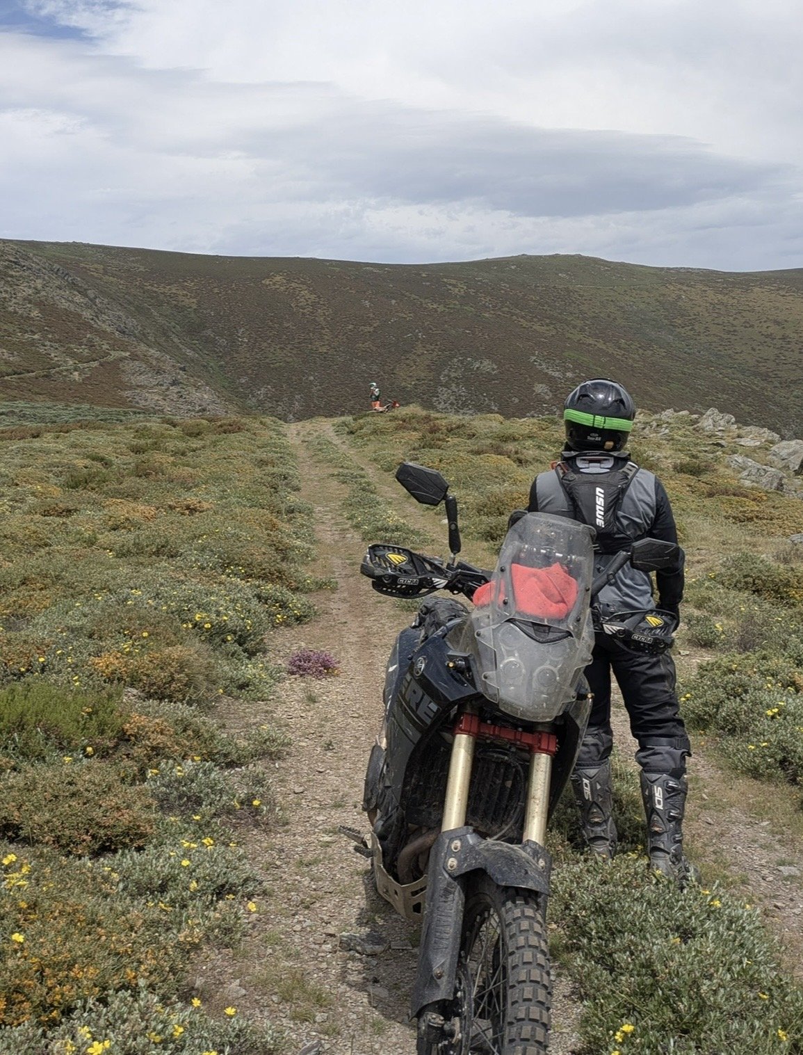 A person dressed in motorcycle gear stands next to a dirt motorcycle on a trail in a hilly, grassy landscape with another rider visible in the distance.