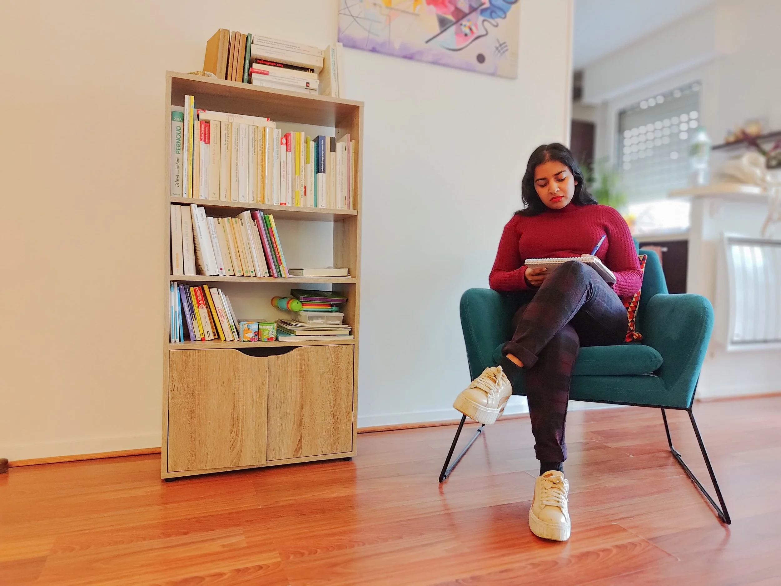 Une jeune femme aux cheveux noirs, portant un pull rouge, des pantalons noirs, et des baskets blanches, assise dans un fauteuil vert en train d'écrire dans un carnet dans un salon lumineux. À côté d'elle, une étagère en bois remplie de livres.