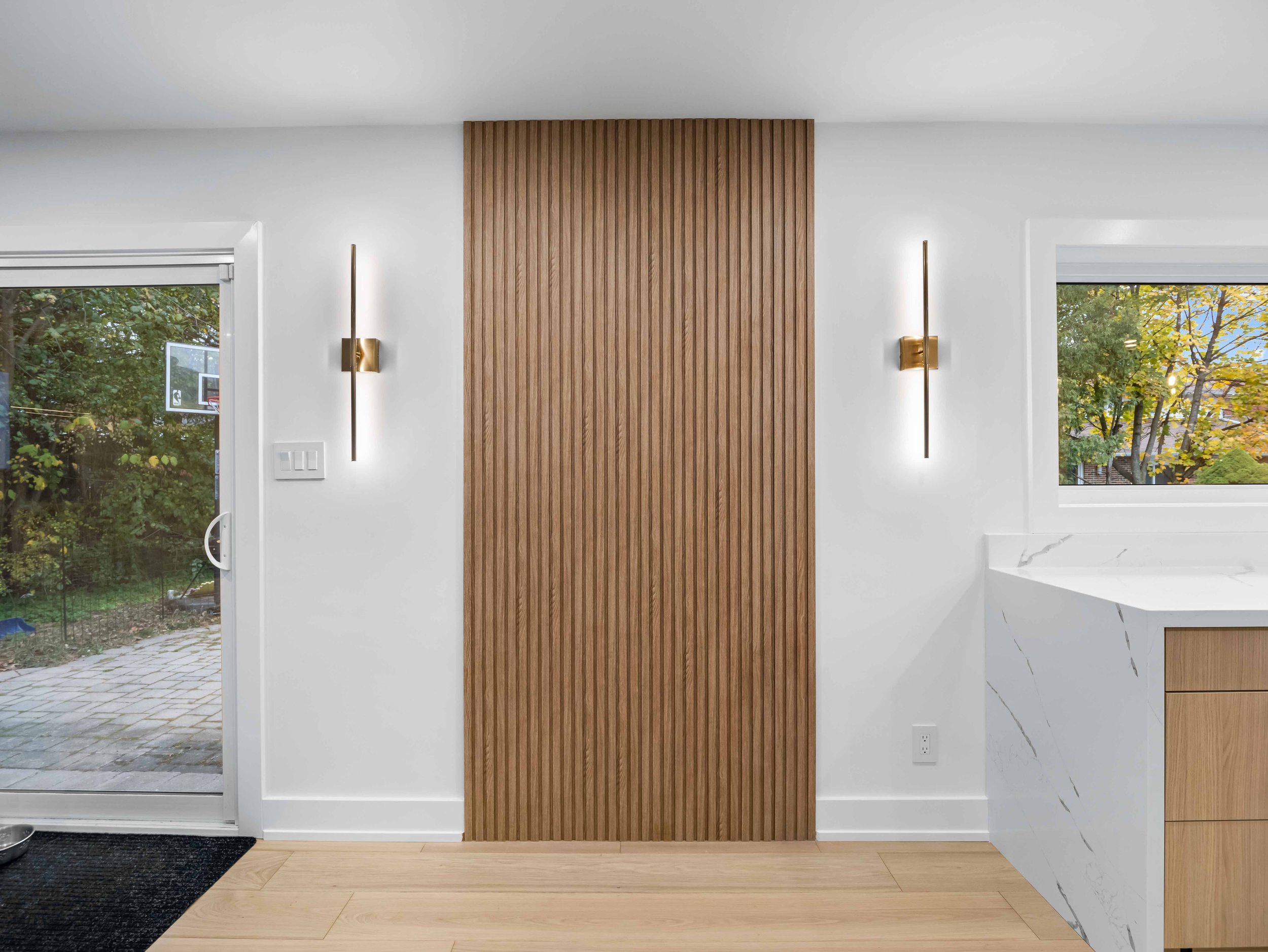 Modern interior space with wooden slat wall, white walls, two vertical wall sconce lights, and two large windows showing green trees outside.