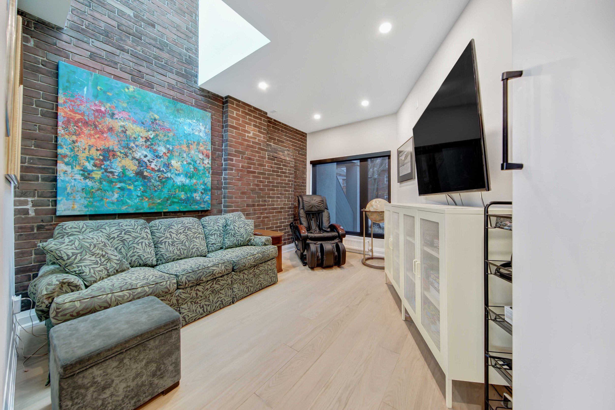 Living room with brick walls, a patterned couch, a black recliner, a large TV mounted on the wall, window with blinds, and abstract artwork on the wall.