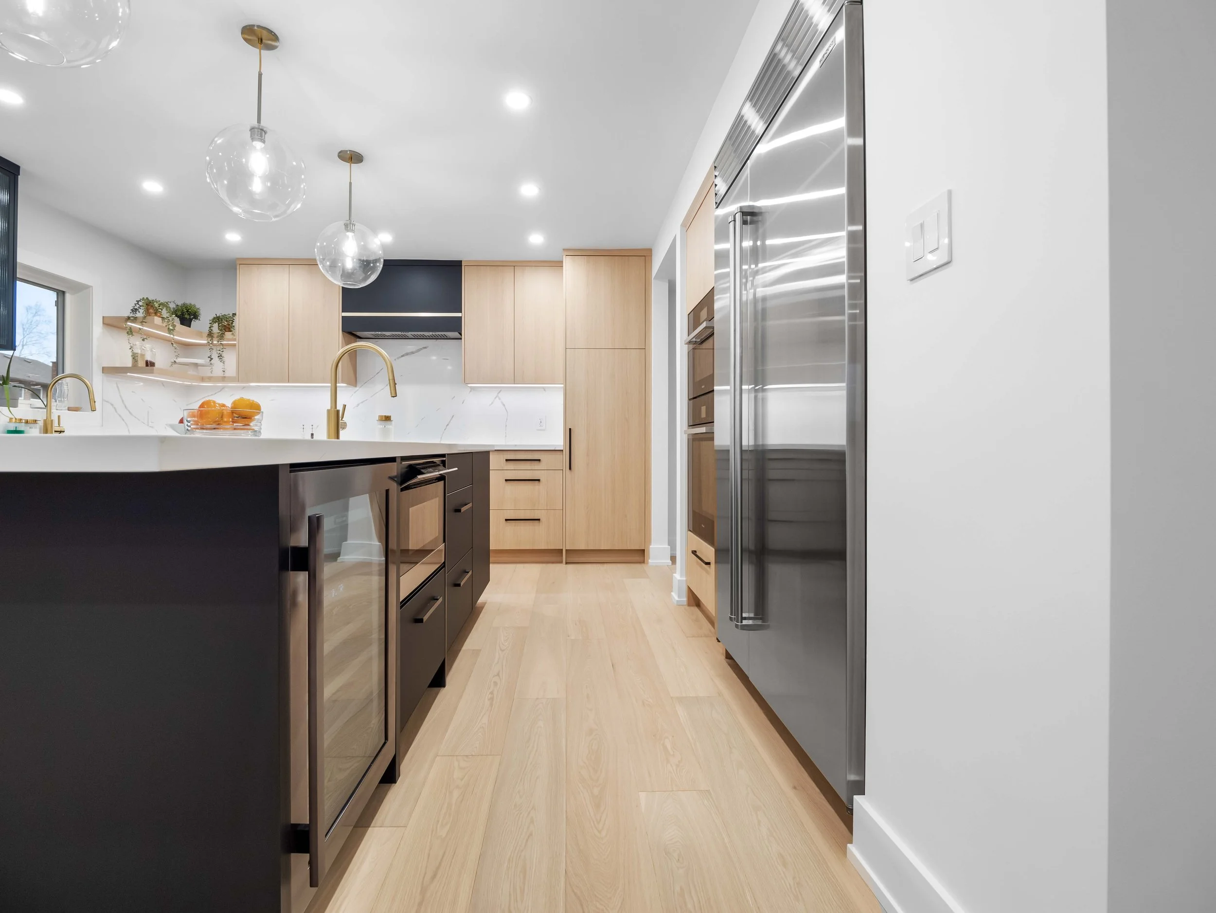 Modern kitchen with light wood cabinets, black lower cabinets, marble backsplash, and stainless steel appliances, including a large refrigerator, with pendant lights hanging from a white ceiling.