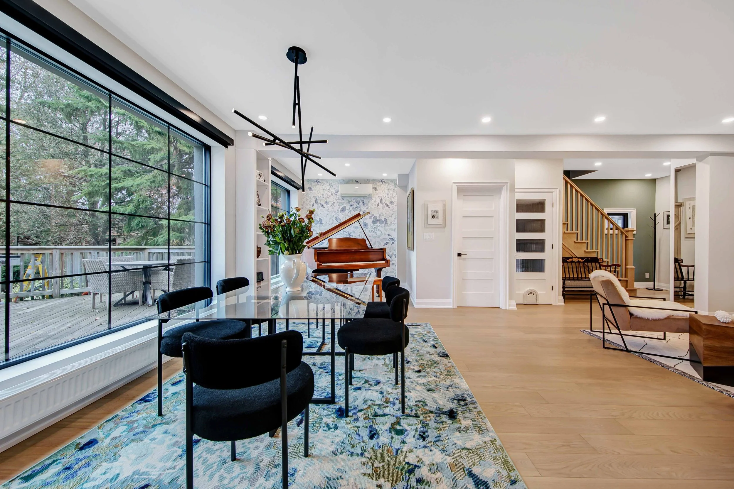 A spacious living room with large windows showing a backyard with trees. The room features a glass dining table with black chairs, a decorative rug, a potted plant, a piano, and a staircase leading upstairs. Modern lighting and artwork adorn the whit
