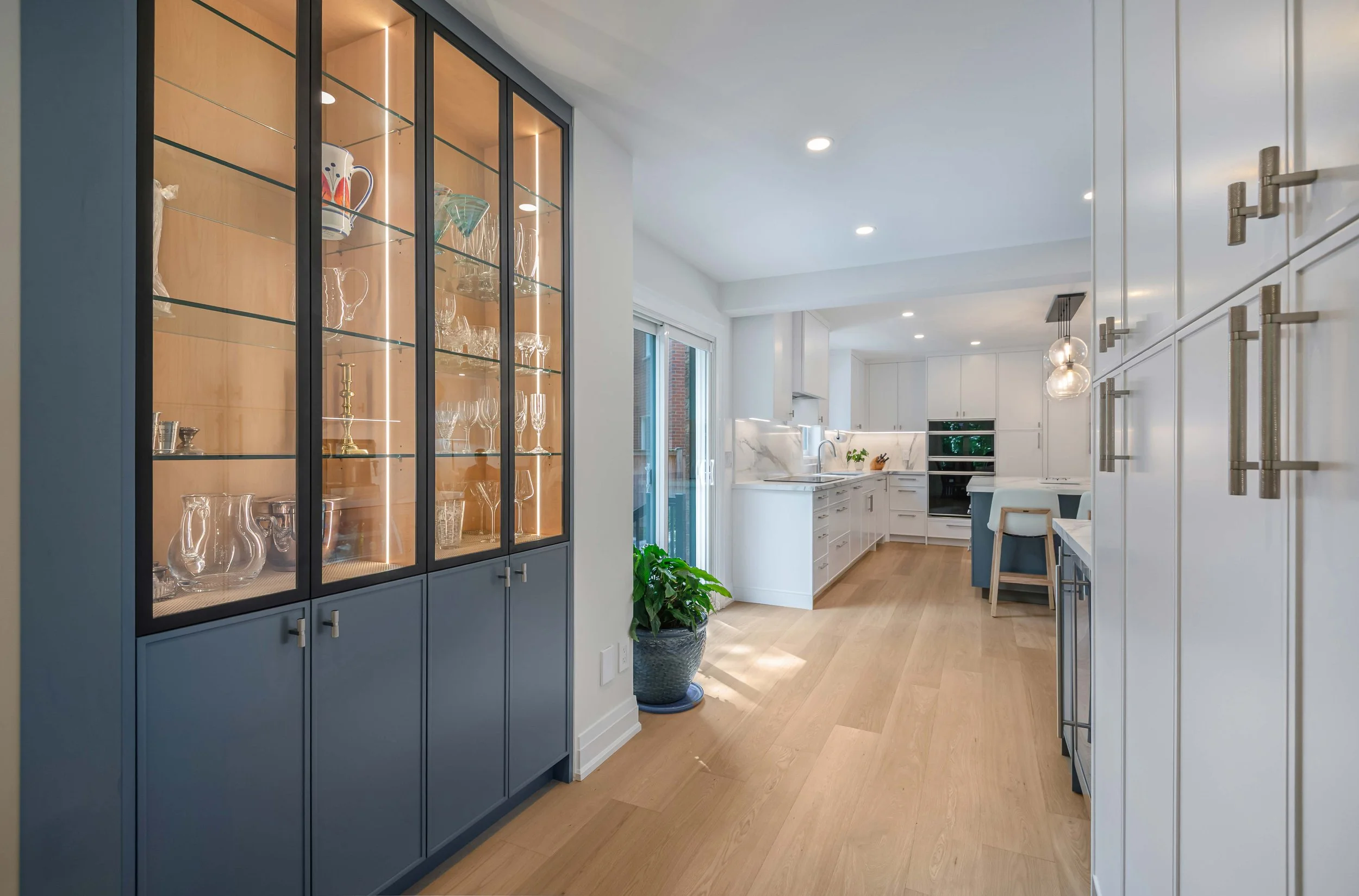 A modern kitchen with white cabinets, a marble backsplash, stainless steel appliances, and a large window. There is a blue display cabinet with glassware, and a green potted plant on the floor near a sliding door leading outside.