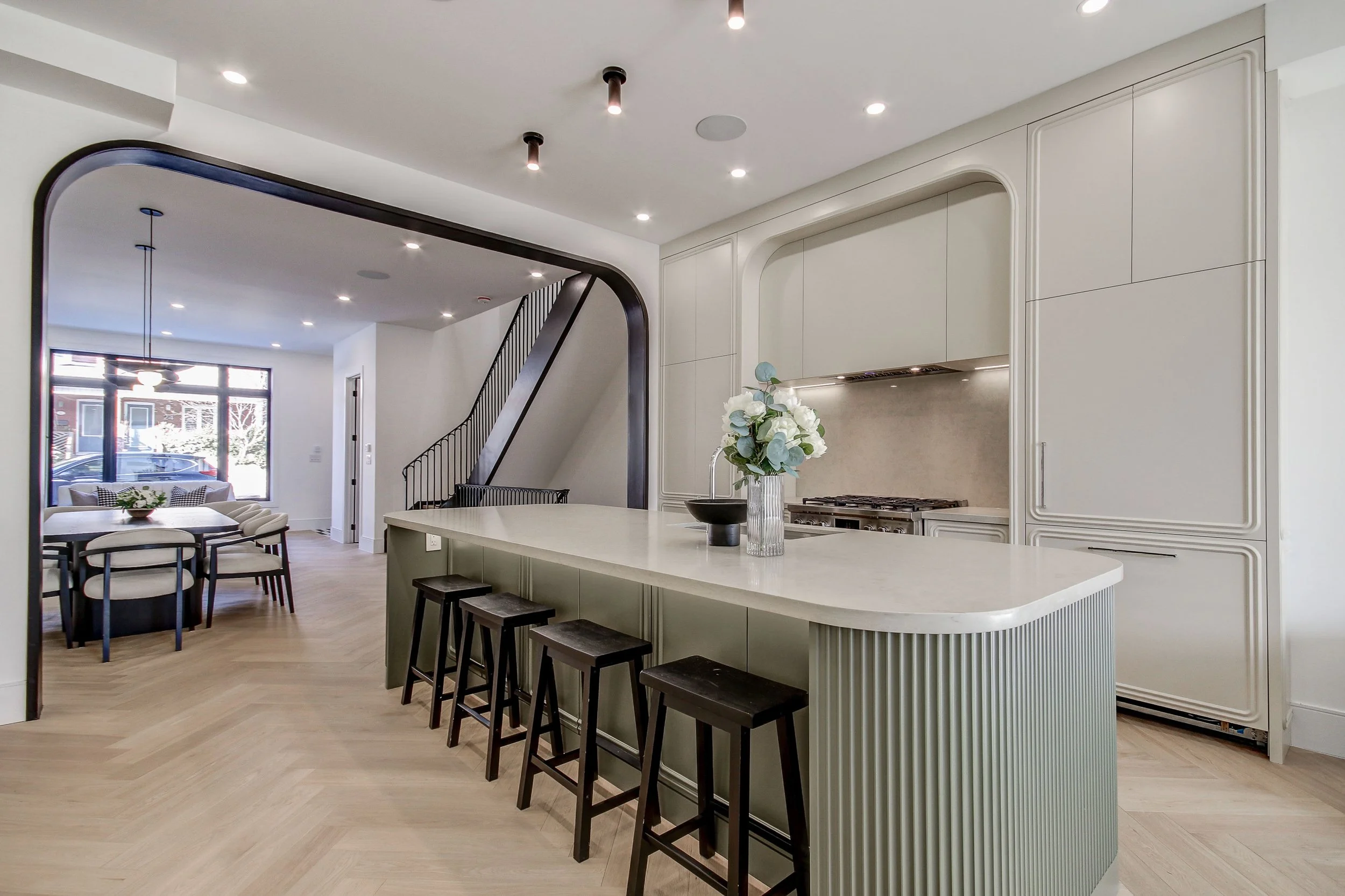 Modern kitchen with white cabinetry, a kitchen island with barstools, and a living area with a dining table and chairs. Large window allowing natural light, light wood flooring, and ceiling lights.