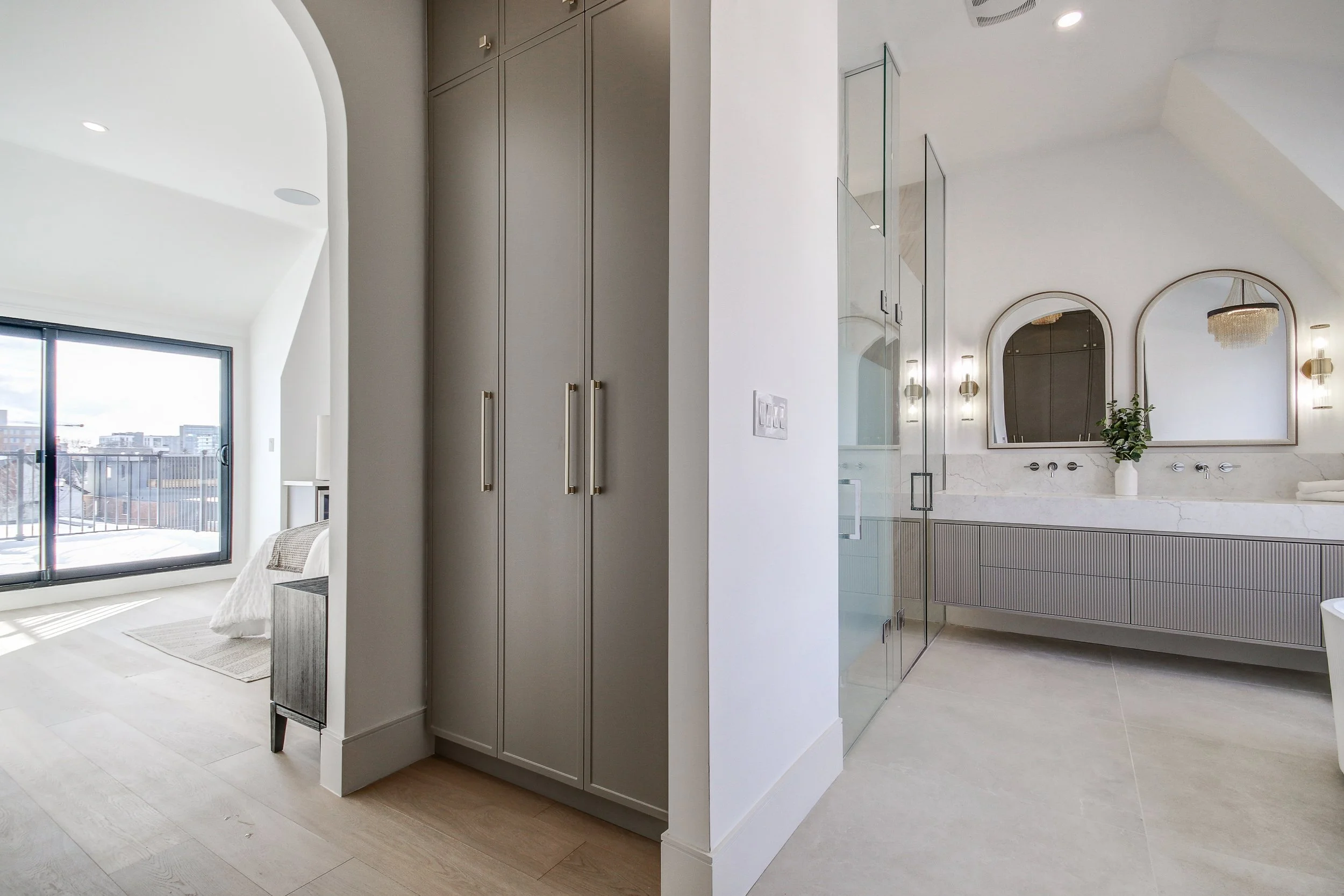 A modern bathroom with white walls, a large mirror, a light fixture, and a vanity with a plant. Adjacent to the bathroom is a bedroom with a bed, nightstand, and a balcony outside the window.