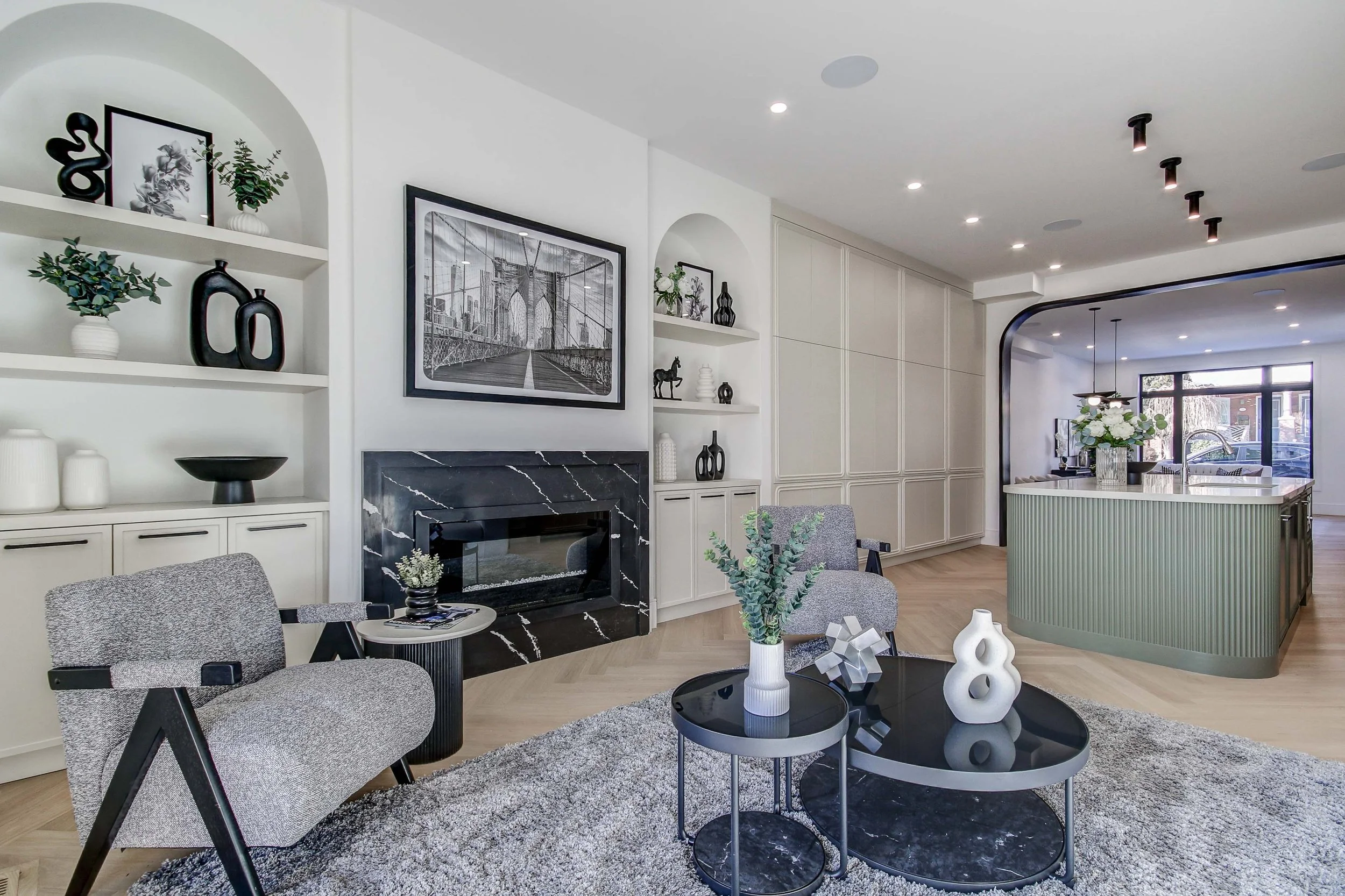 Modern living room with a fireplace, black framed artwork, white built-in shelves decorated with vases and plants, gray armchair, black round coffee table with decorative objects, and a kitchen with a white and green island in the background.
