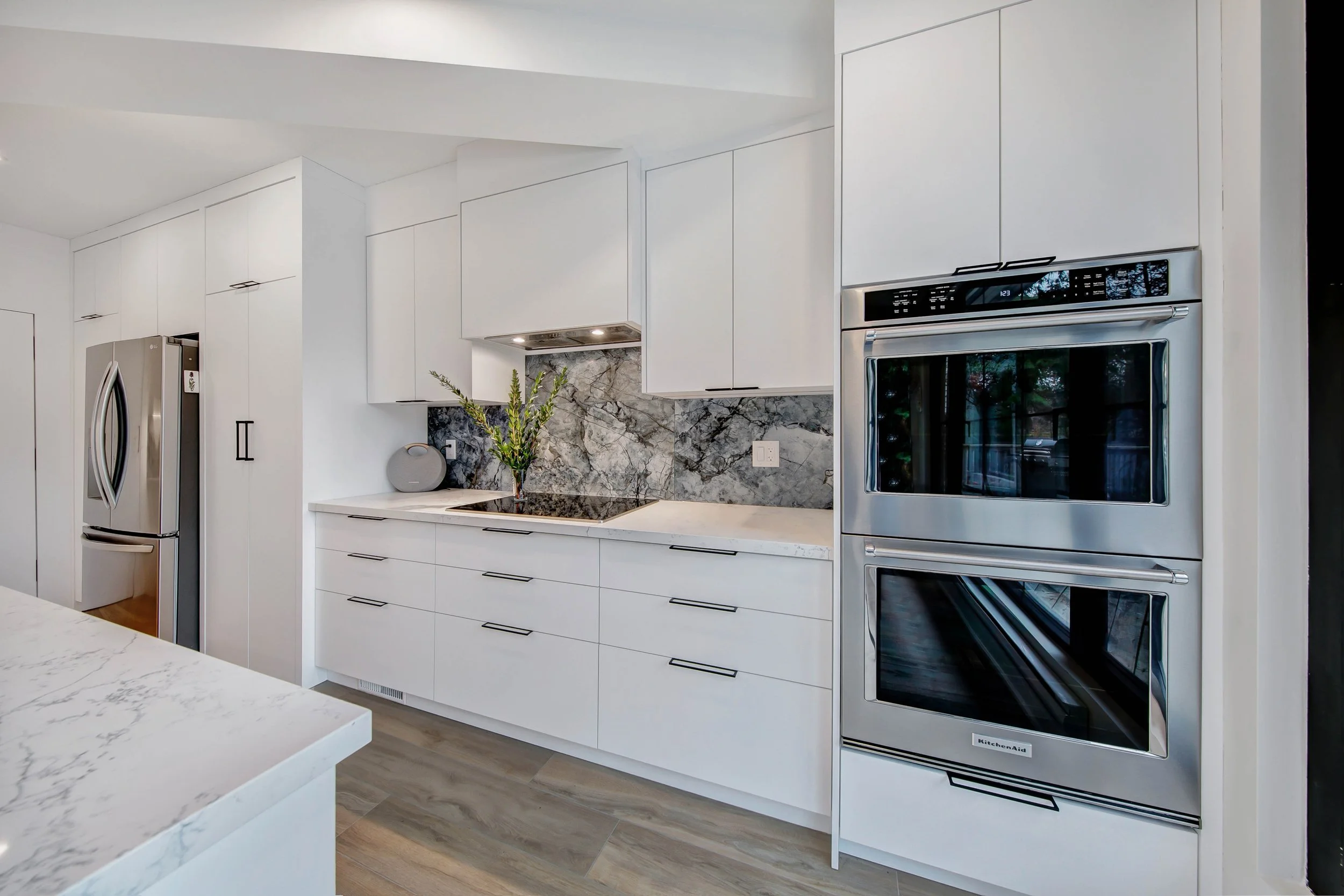 Modern kitchen with white cabinets, a marble backsplash, stainless steel refrigerator, wall oven, and a potted plant on the counter.