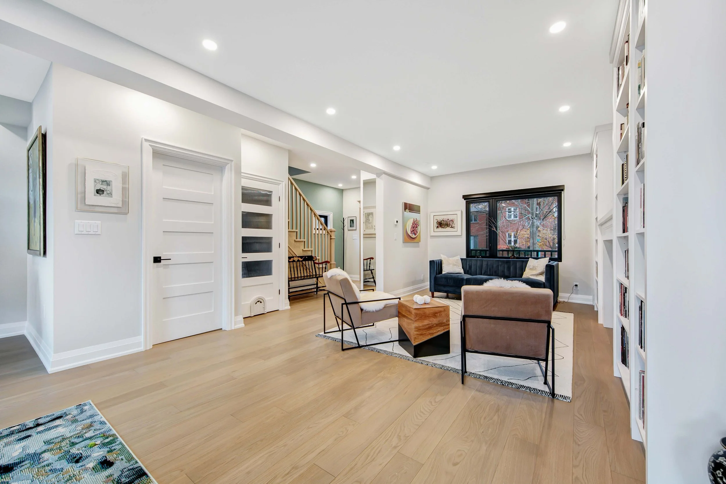Bright living room with white walls and ceiling, hardwood floors, a large window showing trees outside, a black sofa, beige armchairs, a wooden coffee table, and built-in bookshelves filled with books.