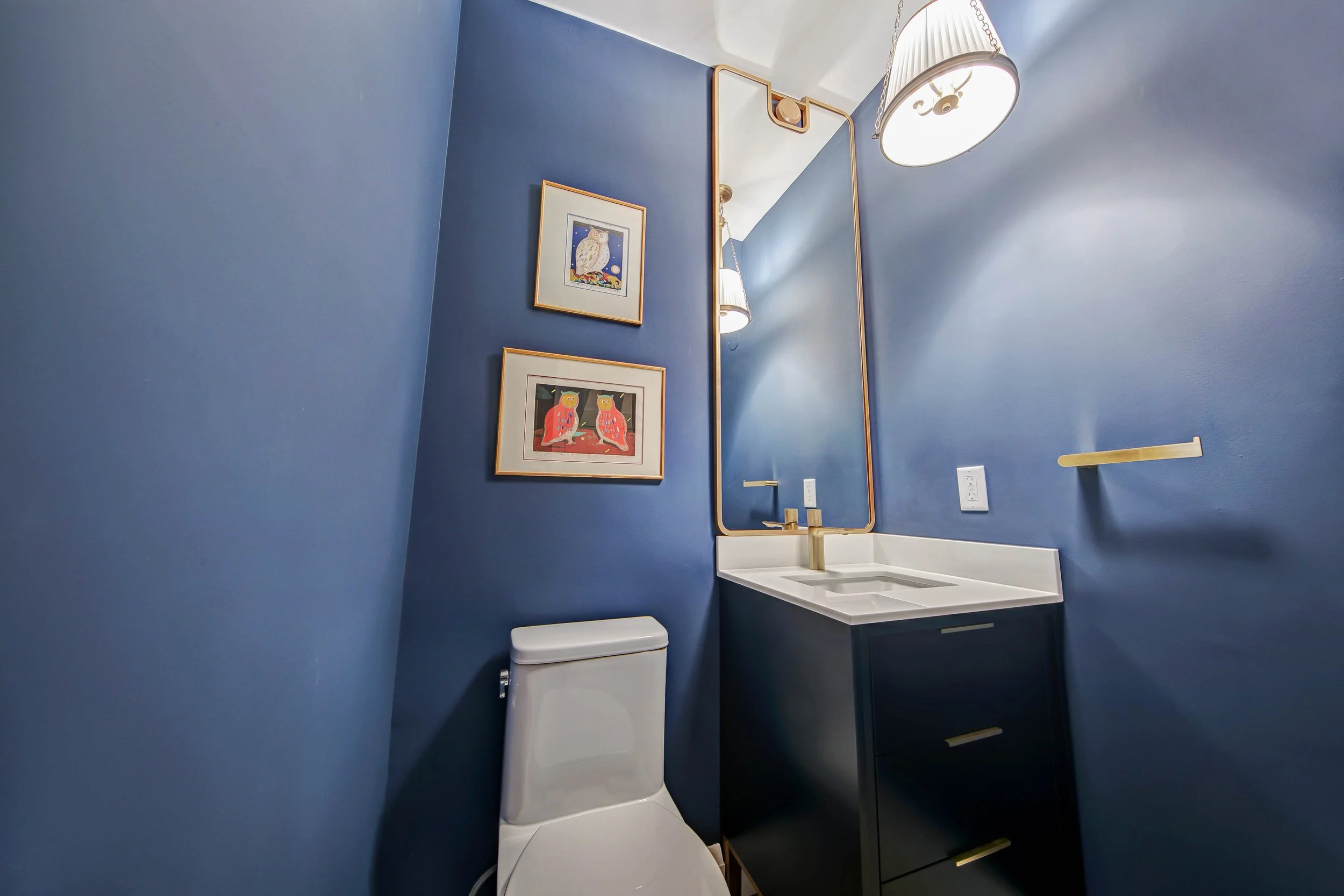 A modern bathroom features blue walls, a white toilet, a white sink with a sleek gold faucet, and black cabinetry with gold handles. There is a large mirror with a gold frame, framed owl and bird artwork, a gold towel bar, and two wall-mounted lamps 