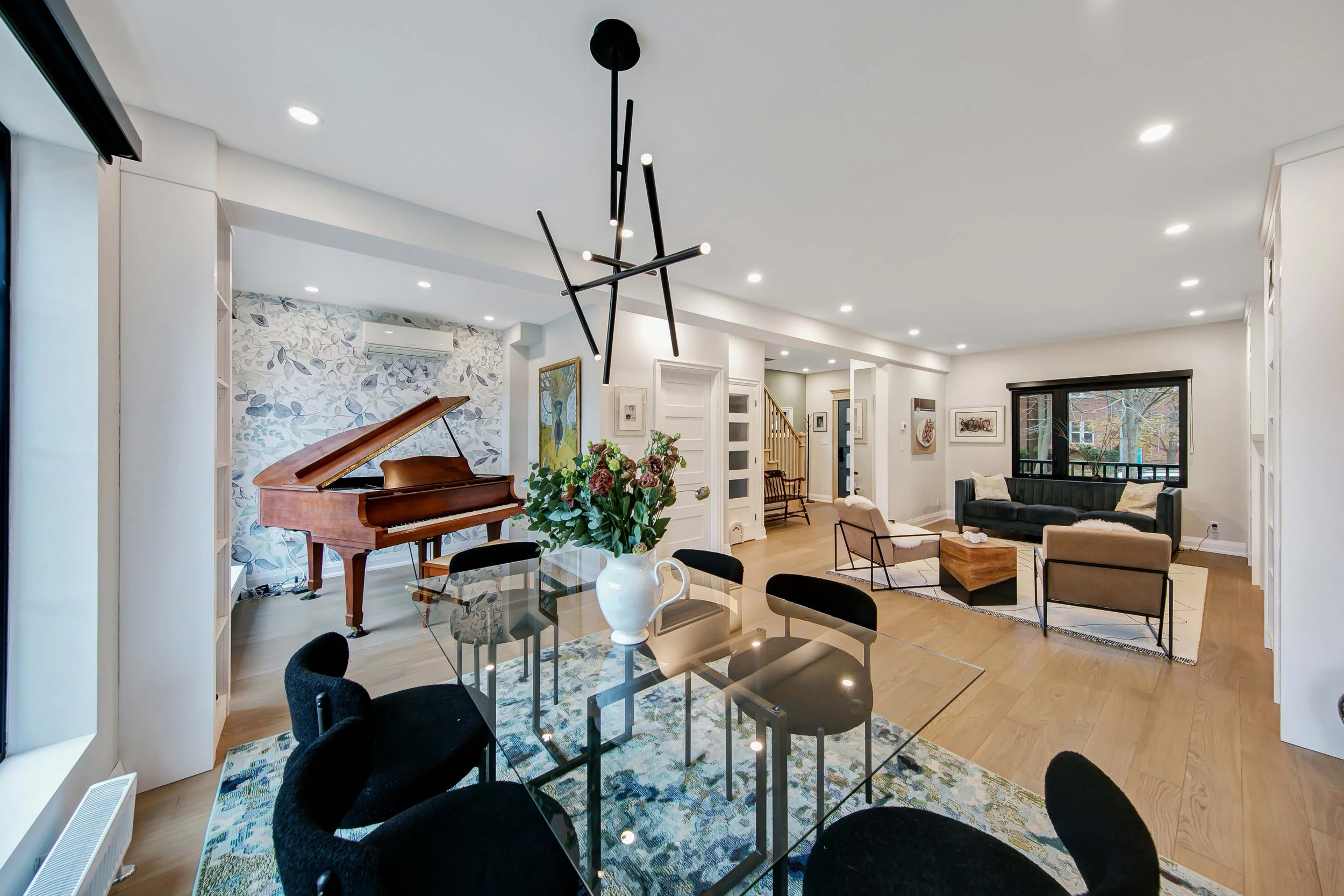 Living room with a grand piano, glass dining table with black chairs, modern chandelier, sofa area, artwork, large windows, hardwood floors, and white walls.