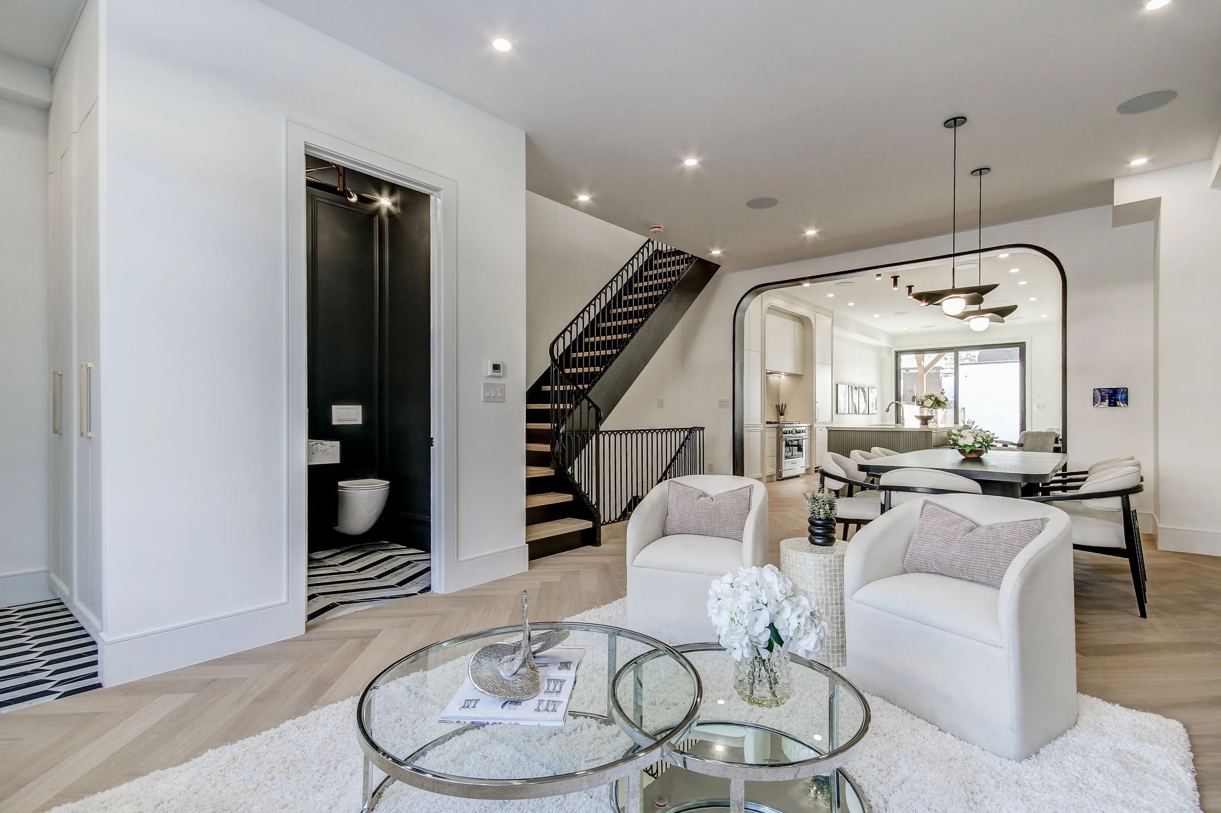 Modern living room with white walls and ceiling, featuring a staircase with black railing, a round glass coffee table on a white shag rug, white armchairs, and a dining area with a table and chairs near large windows and a kitchenette.