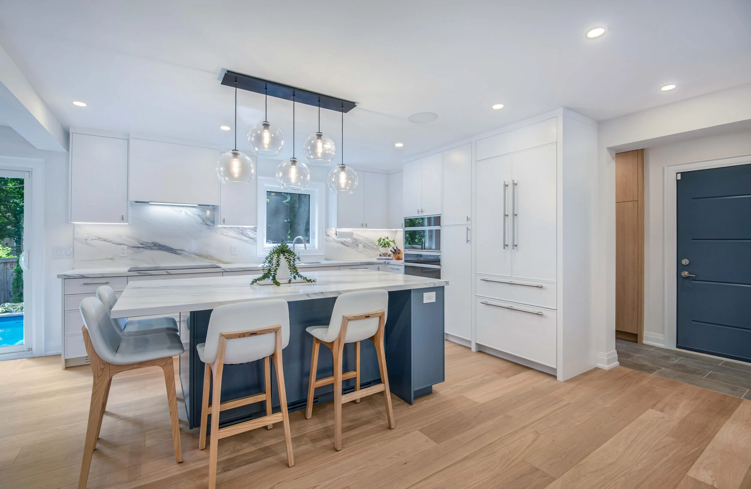 Modern white kitchen with a marble backsplash, pendant lights hanging over a kitchen island with blue base, surrounded by white chairs with wooden legs, and wooden floors.