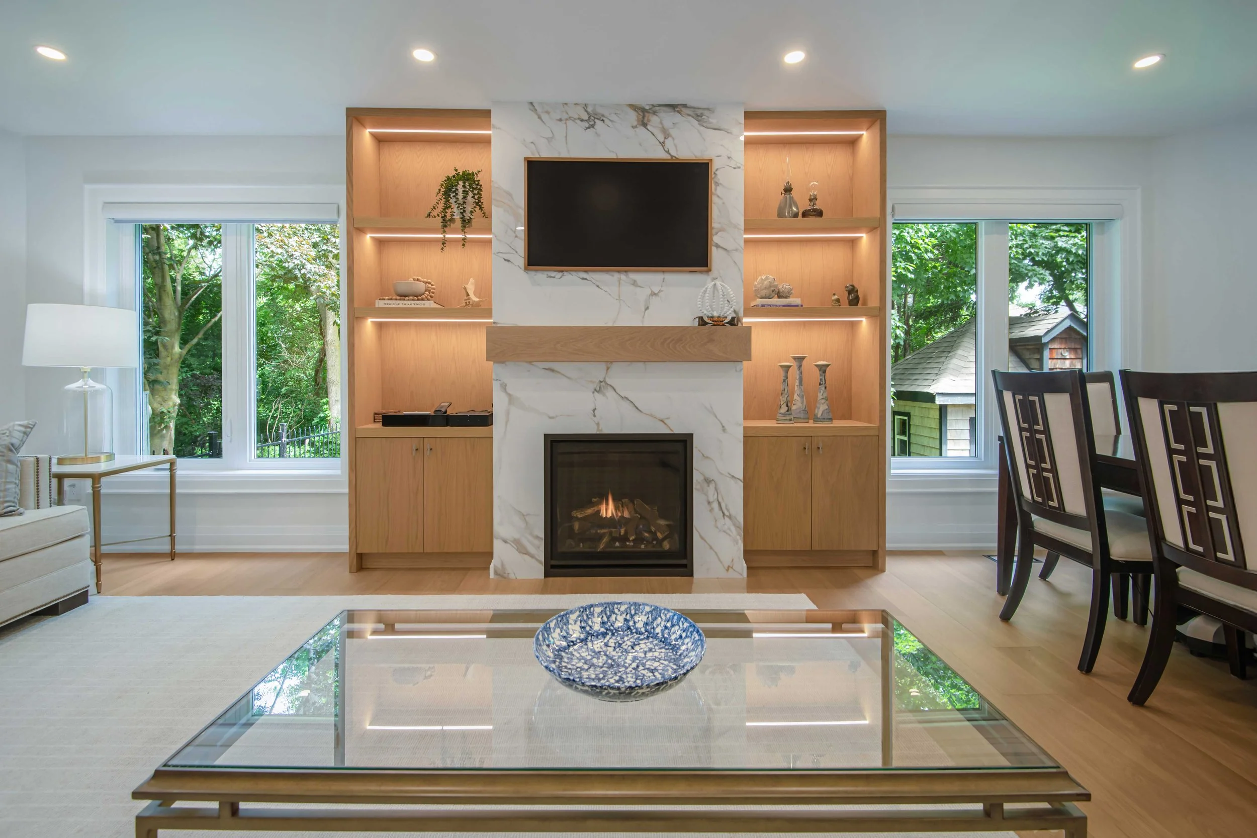 Living room with a modern fireplace, built-in wooden shelves with decorative items, two windows showing green trees, a dining table with chairs, and a glass coffee table with a blue decorative bowl.