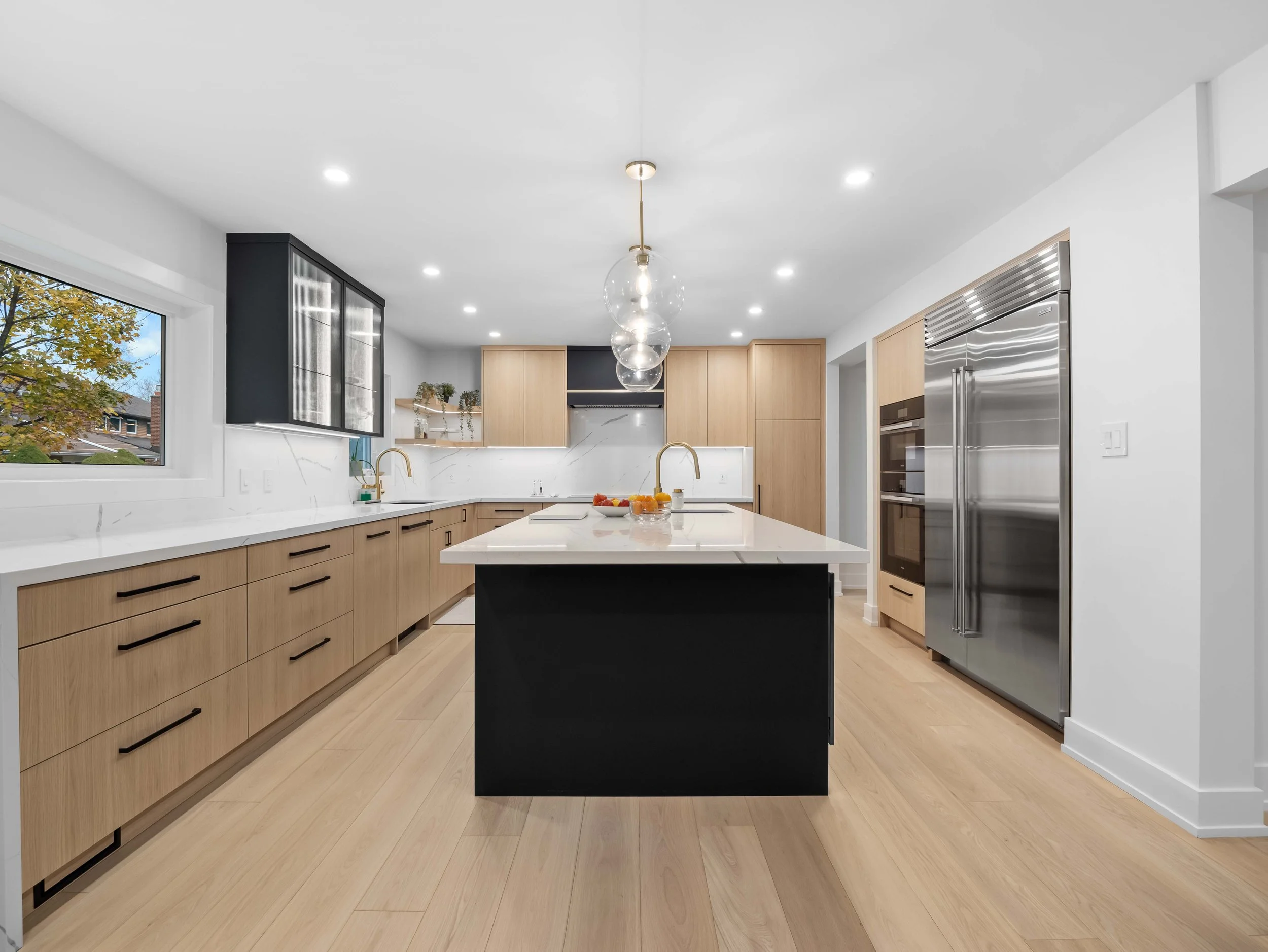 Modern kitchen with light wood cabinets, a white island with a black base, a stainless steel refrigerator, and a window overlooking trees.