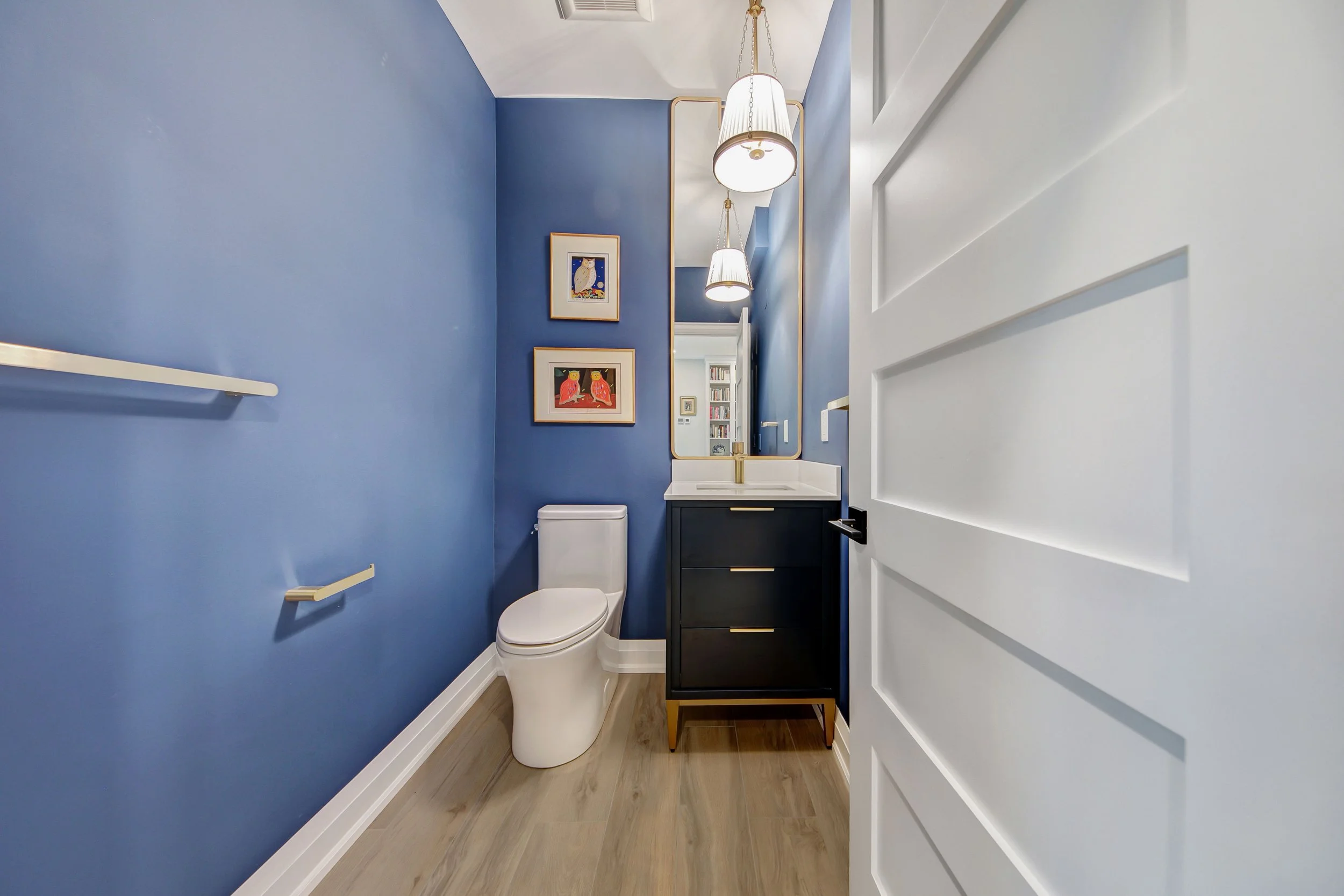 A small bathroom with a white door, light wood flooring, and blue walls. The bathroom features a modern white toilet, a black vanity with a gold handle, a rectangular mirror with a gold frame, two framed owl artwork on the wall, and two hanging penda