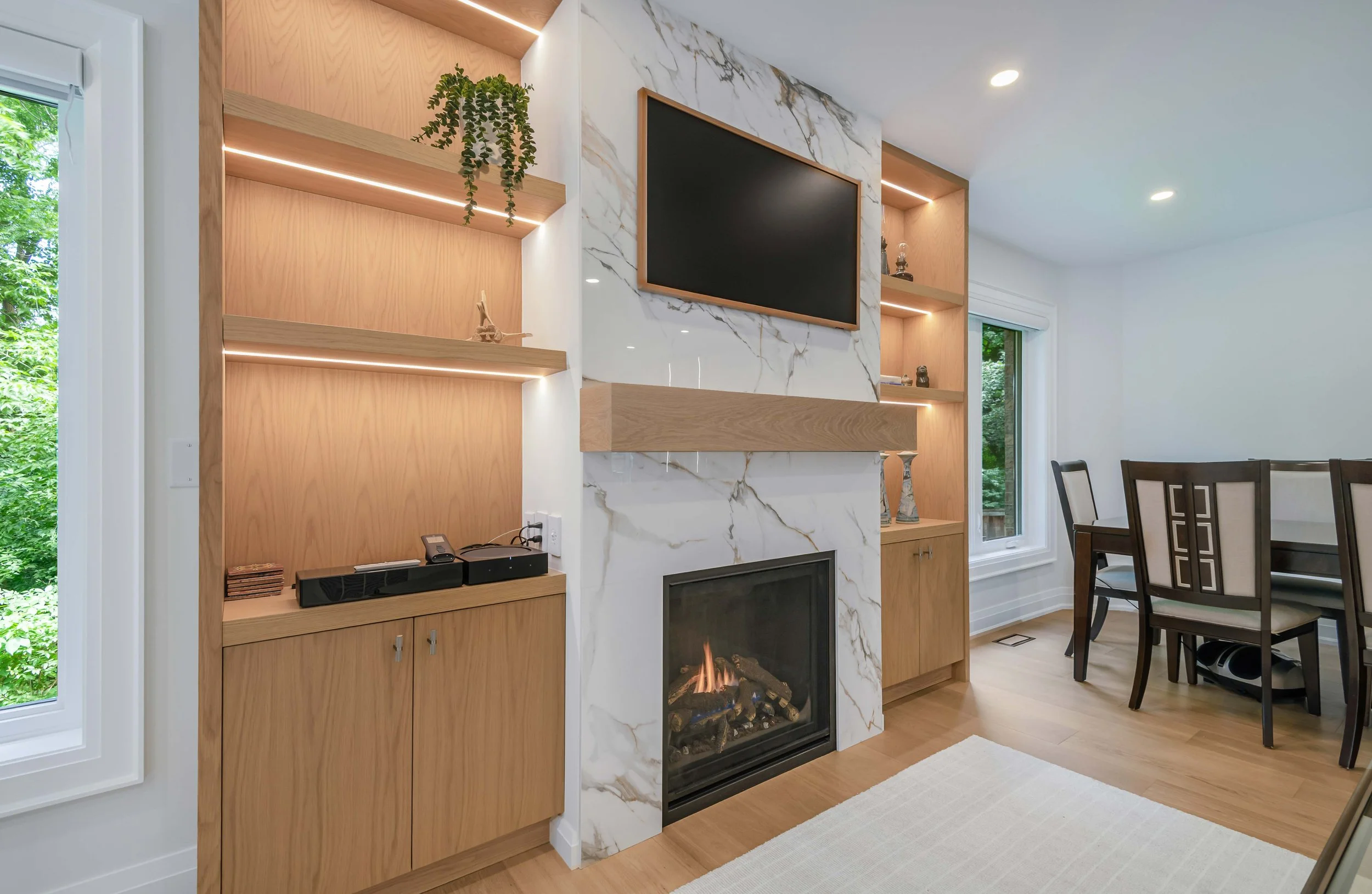 Living room with a fireplace framed by white marble with gold veining, adjacent to built-in wooden shelves and cabinetry, a wall-mounted flat-screen TV, and large windows with green foliage outside.