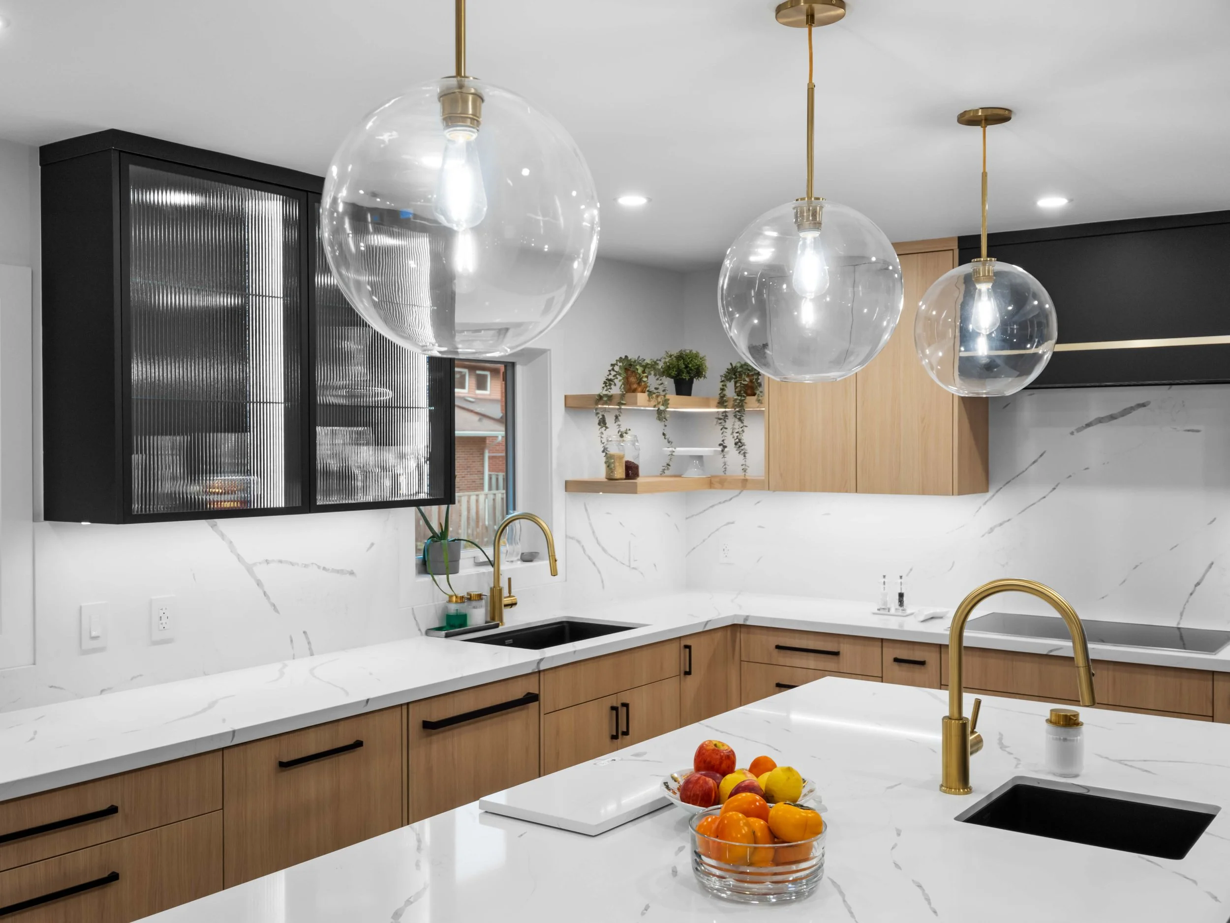 Modern kitchen with a white marble counter, wooden cabinets, and three large glass pendant lights hanging from the ceiling. A bowl of mixed fruit is on the counter near the sink.