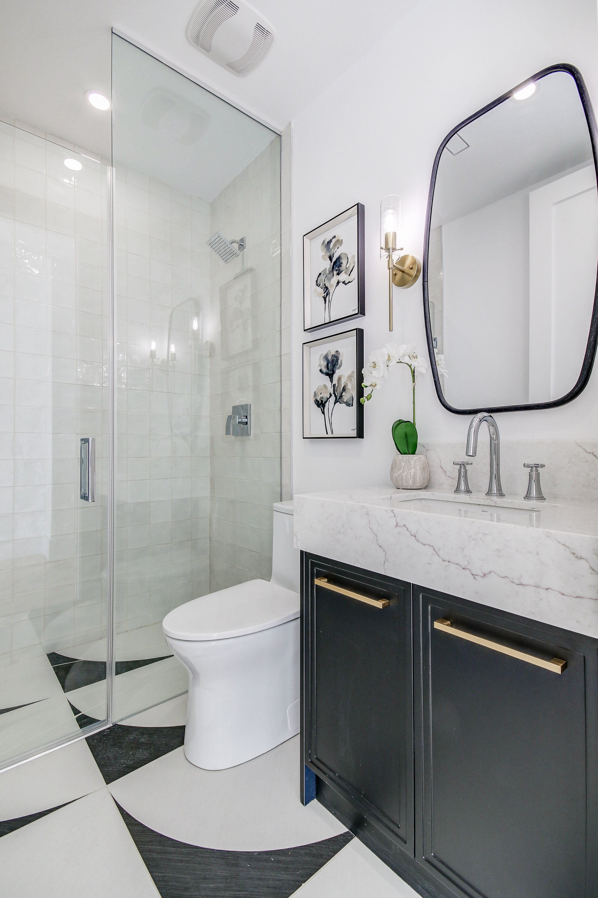 Modern bathroom with a walk-in shower with glass door, white toilet, marble vanity with dark cabinet, mirror, two framed floral prints, wall sconce, and decorative black and white tiled floor.