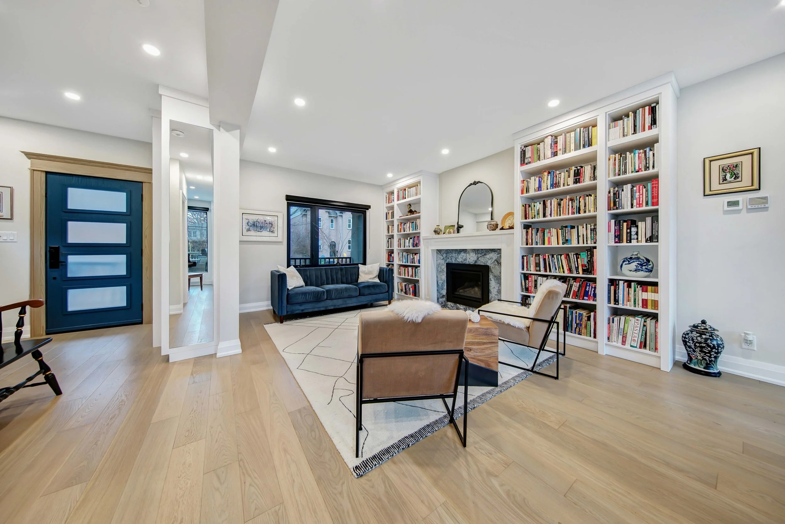 A spacious living room with a white ceiling, hardwood floors, white walls, and recessed lighting. There is a blue front door, a window with black trim, and built-in white bookshelves filled with books. The seating includes a dark blue sofa, beige arm