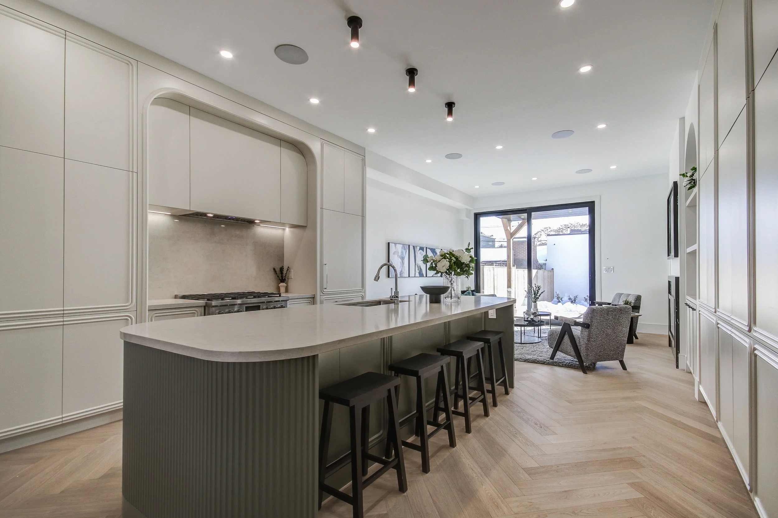 A modern open-concept kitchen and living room with white cabinetry, a large kitchen island with barstools, a dining area with a vase of white flowers, a sitting area with armchairs, and sliding glass doors leading to an outdoor space.