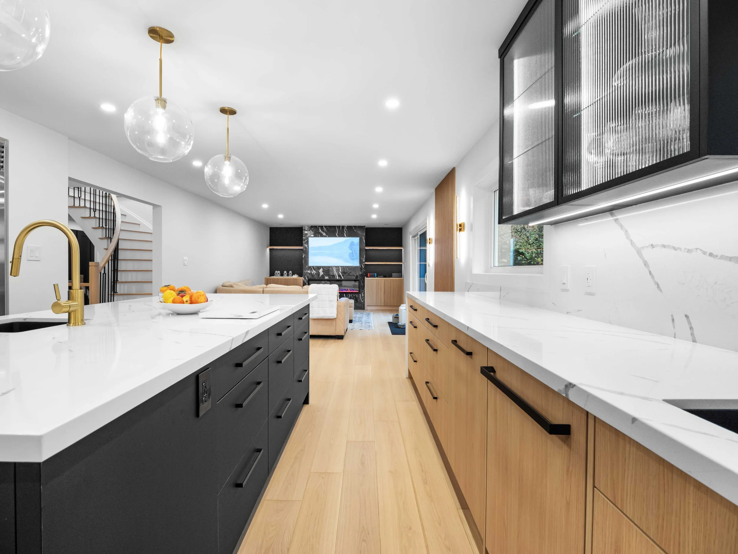 A modern kitchen with a white marble island, black and wood cabinets, hanging glass pendant lights, and a view into a living room with a TV and staircase.