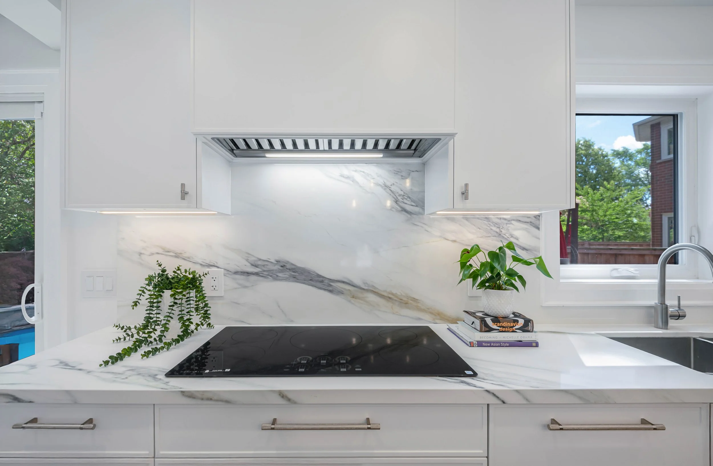 Modern kitchen with white cabinetry, marble backsplash, black stovetop, and open book display with plants on white countertop.